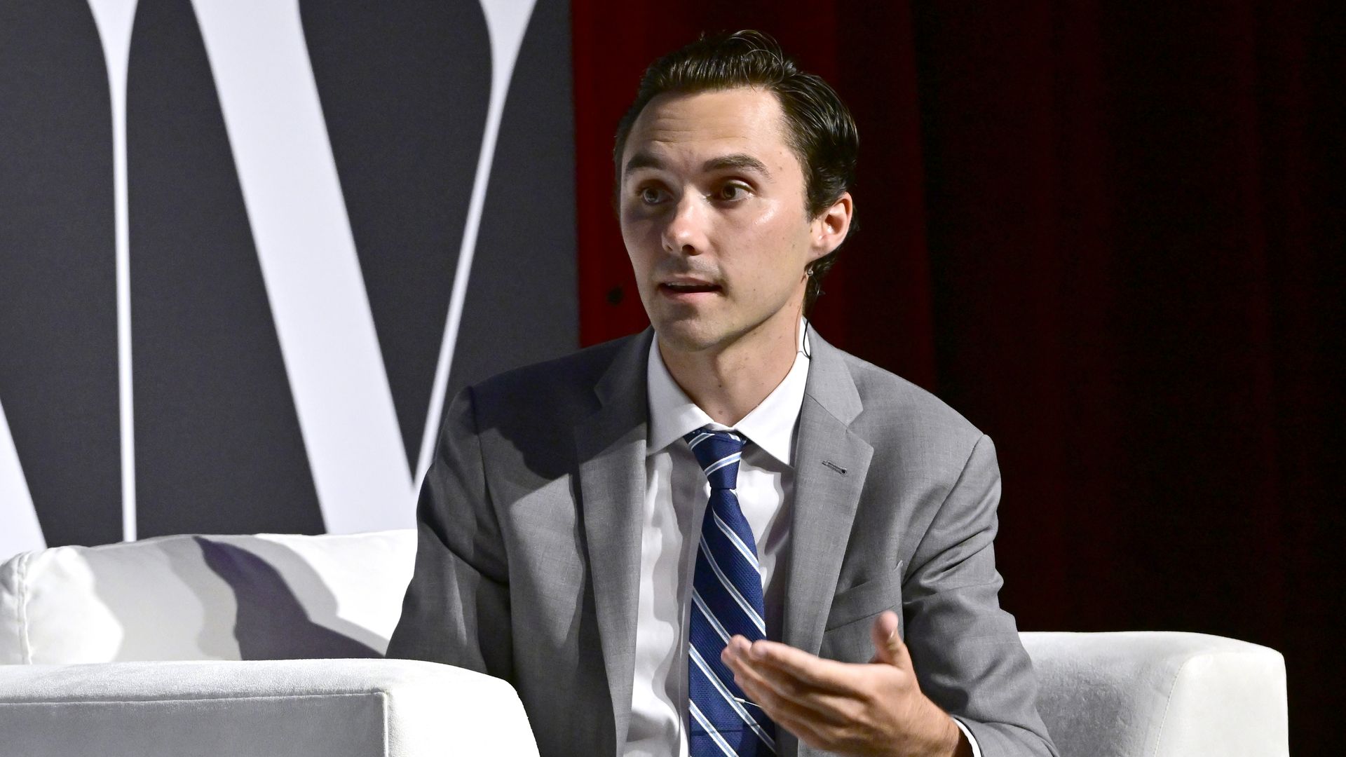 David Hogg sits in a white chair wearing a gray suit in front of a gray and red backdrop.