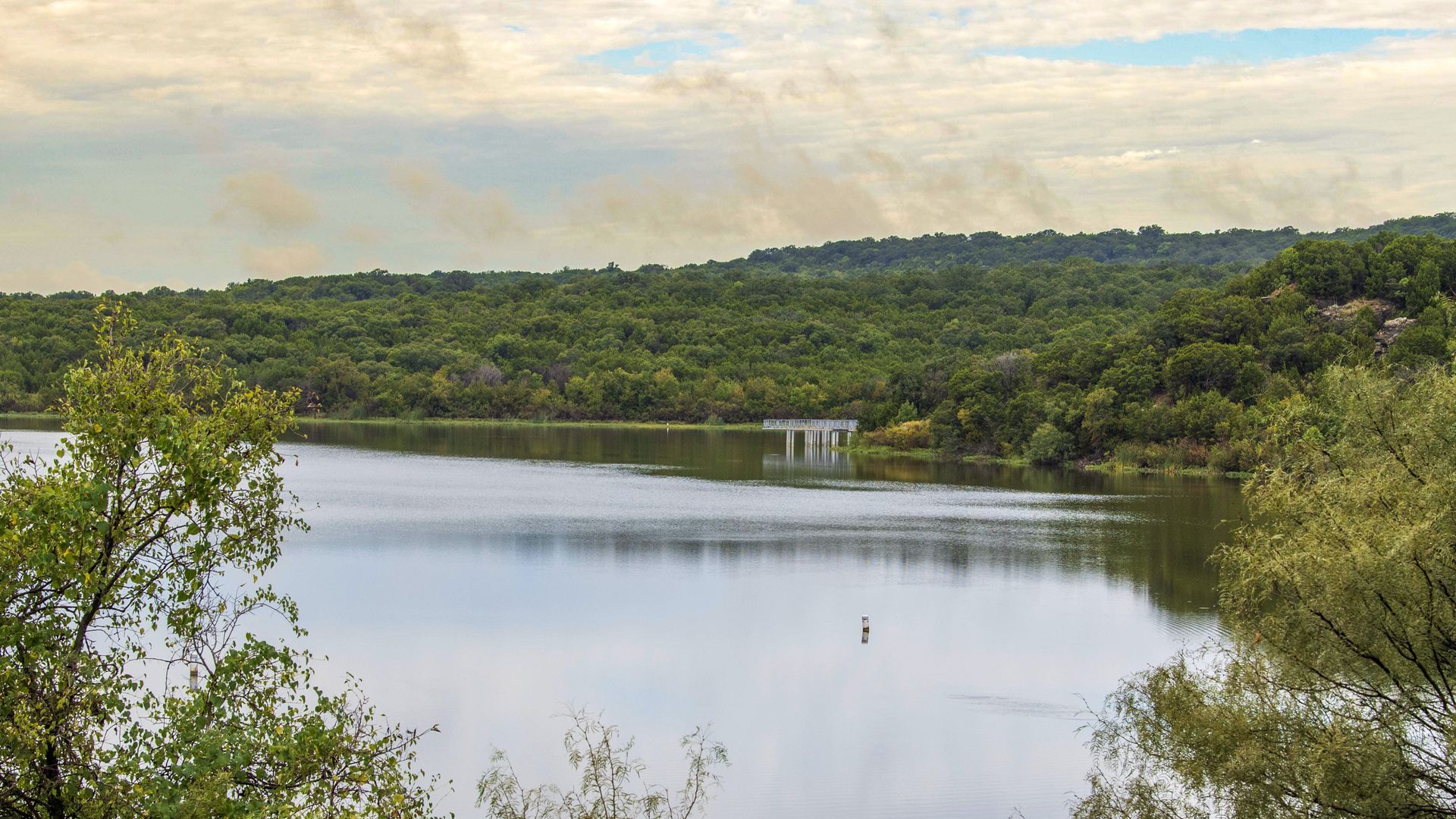 Calm lake surrounded by dense green trees with hills in the background and a partly cloudy sky above. A small pier extends into the water near the forested shore.