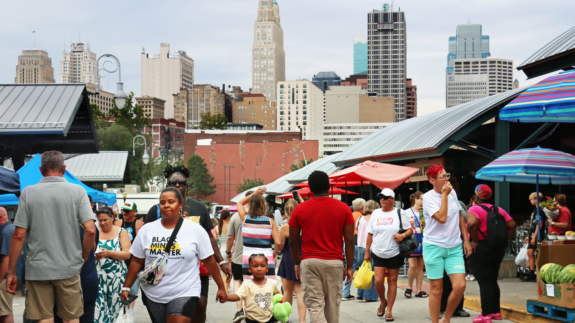 Busy outdoor market scene with diverse people walking along stalls with colorful umbrellas, tall buildings in the background under a cloudy sky.
