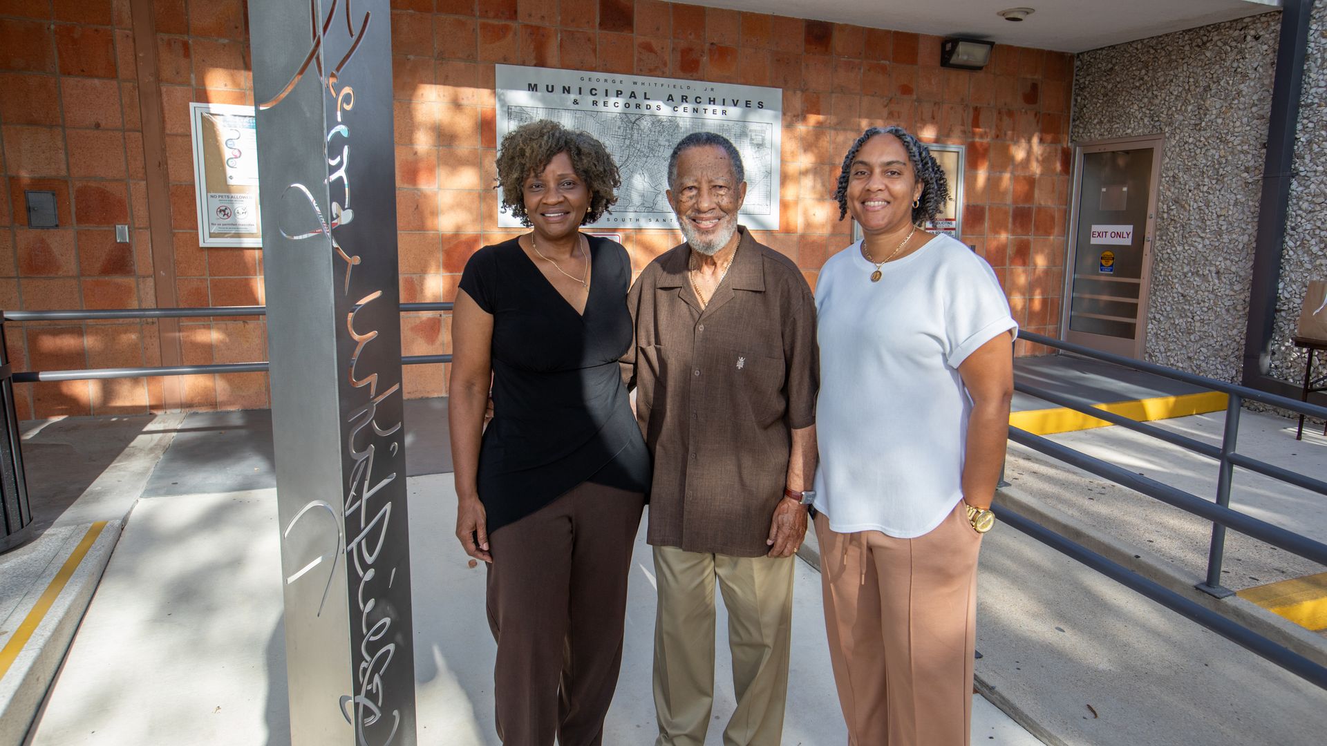 Three smiling adults stand outside a brick Municipal Archives & Records Center building; behind them a sign reads "George Whitfield Jr Municipal Archives & Records Center".