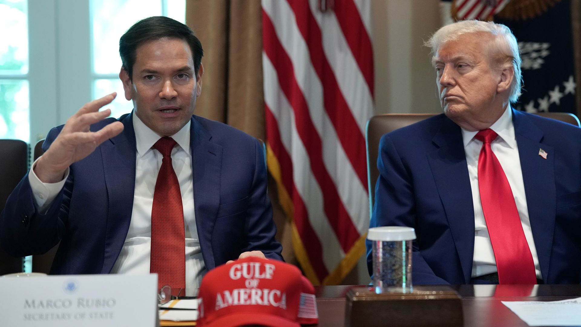 Marco Rubio speaks at a Cabinet meeting while President Trump looks on.  A red Gulf of America hat sits on the table.