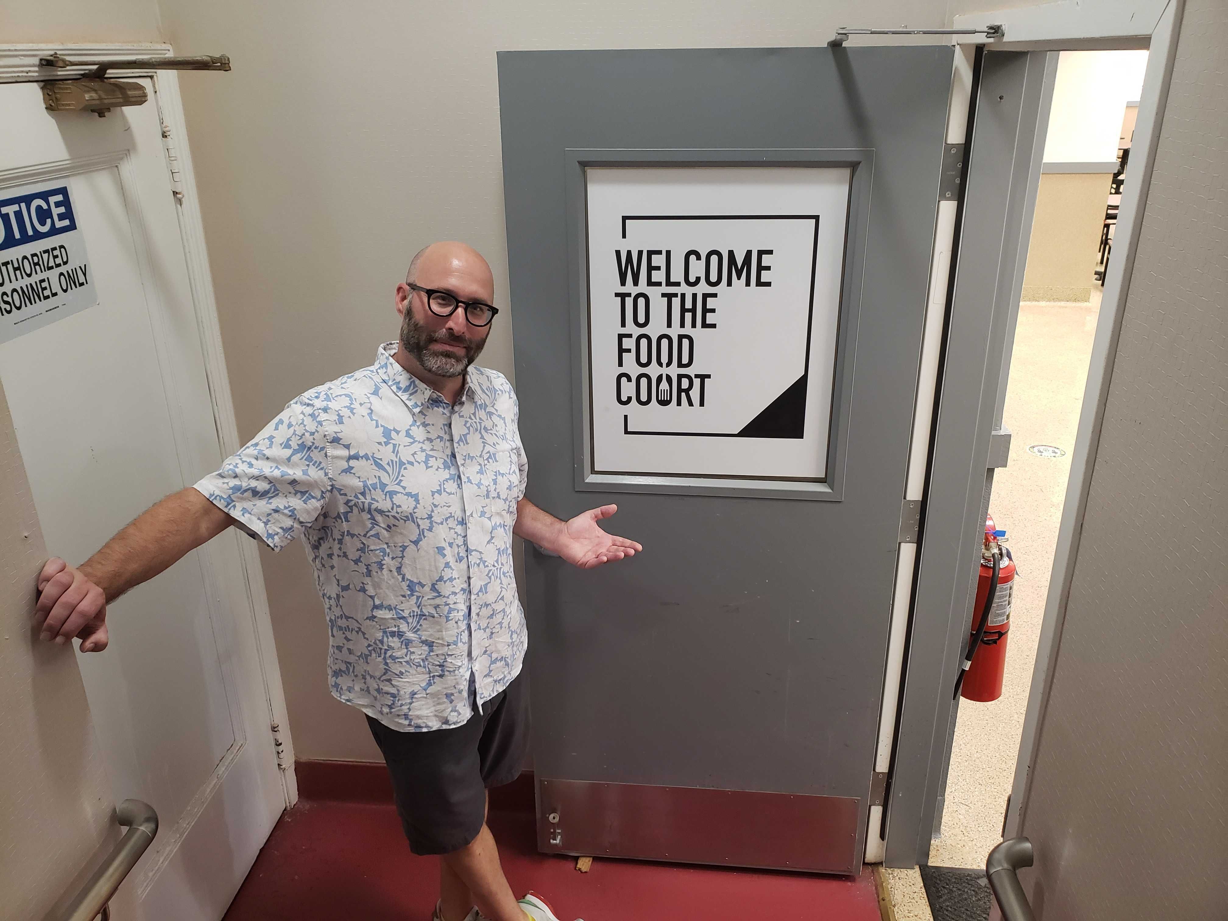 Man pointing to sign that reads 'Welcome to the Food Court' on a gray door.