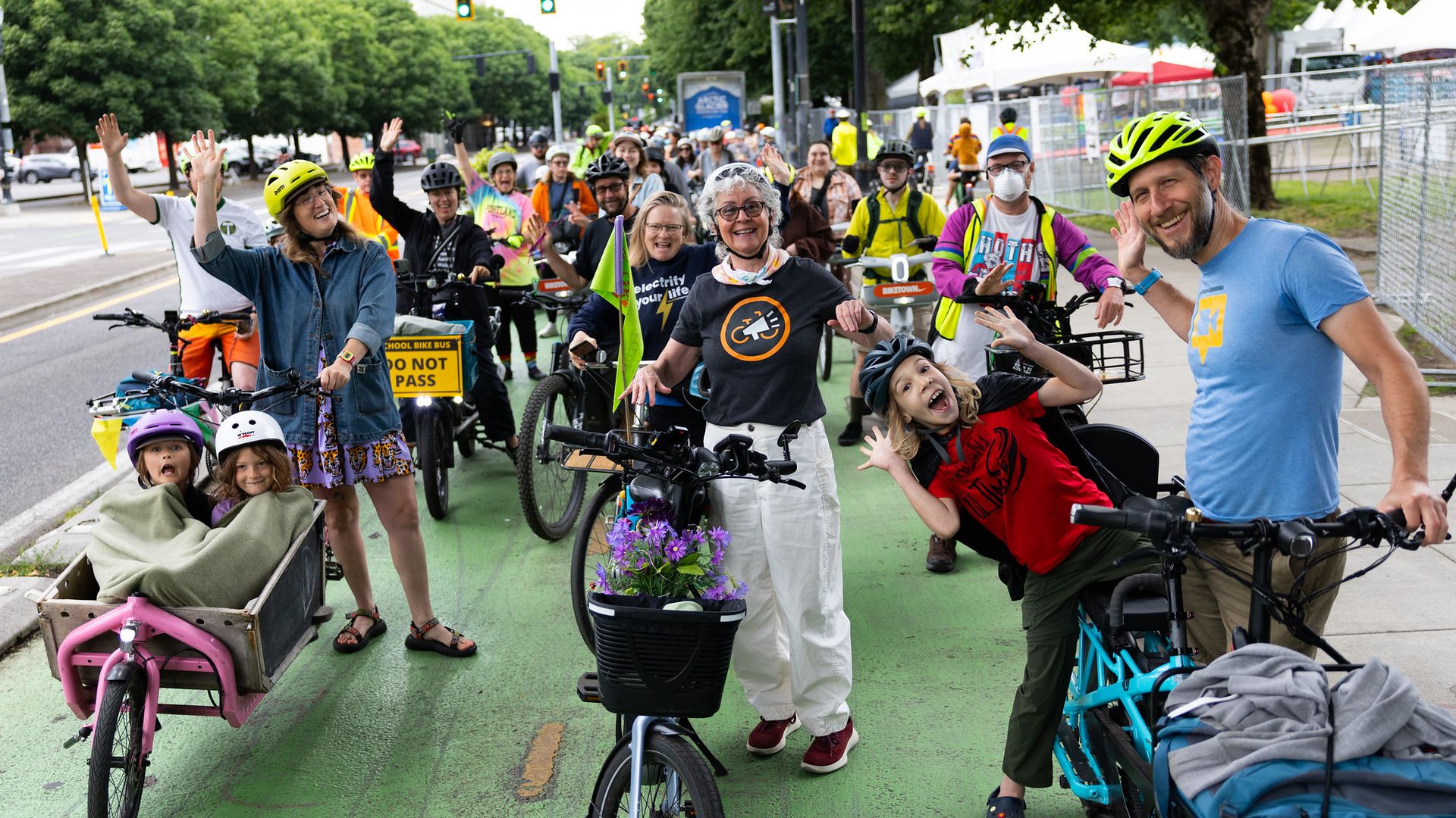 Large group of cyclists with bright helmets wave and smile on a closed city street with a green bike lane, white tents, and bystanders; two kids ride in a wagon on the left.