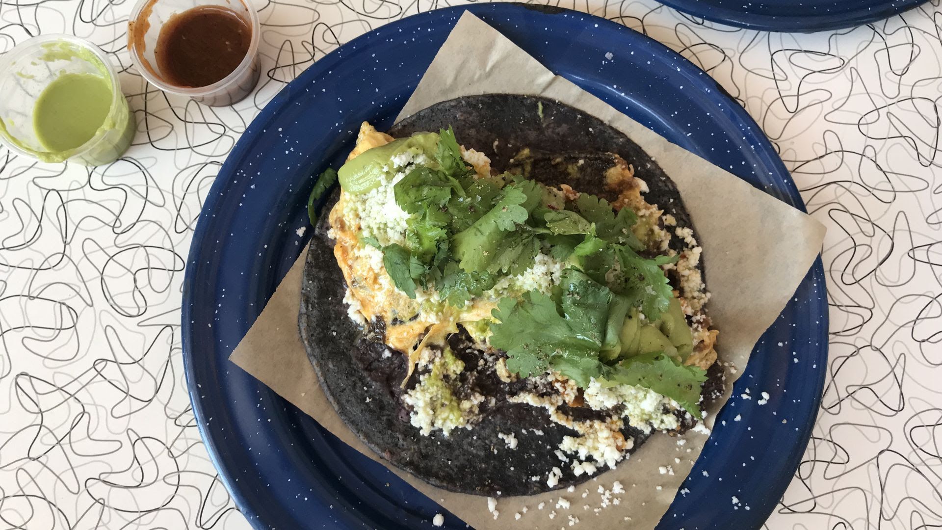 Top-down view of a dark tortilla tostada topped with avocado, cilantro, and crumbled cheese on parchment; blue plate, abstract patterned table, with green salsa and brown sauce cups nearby.