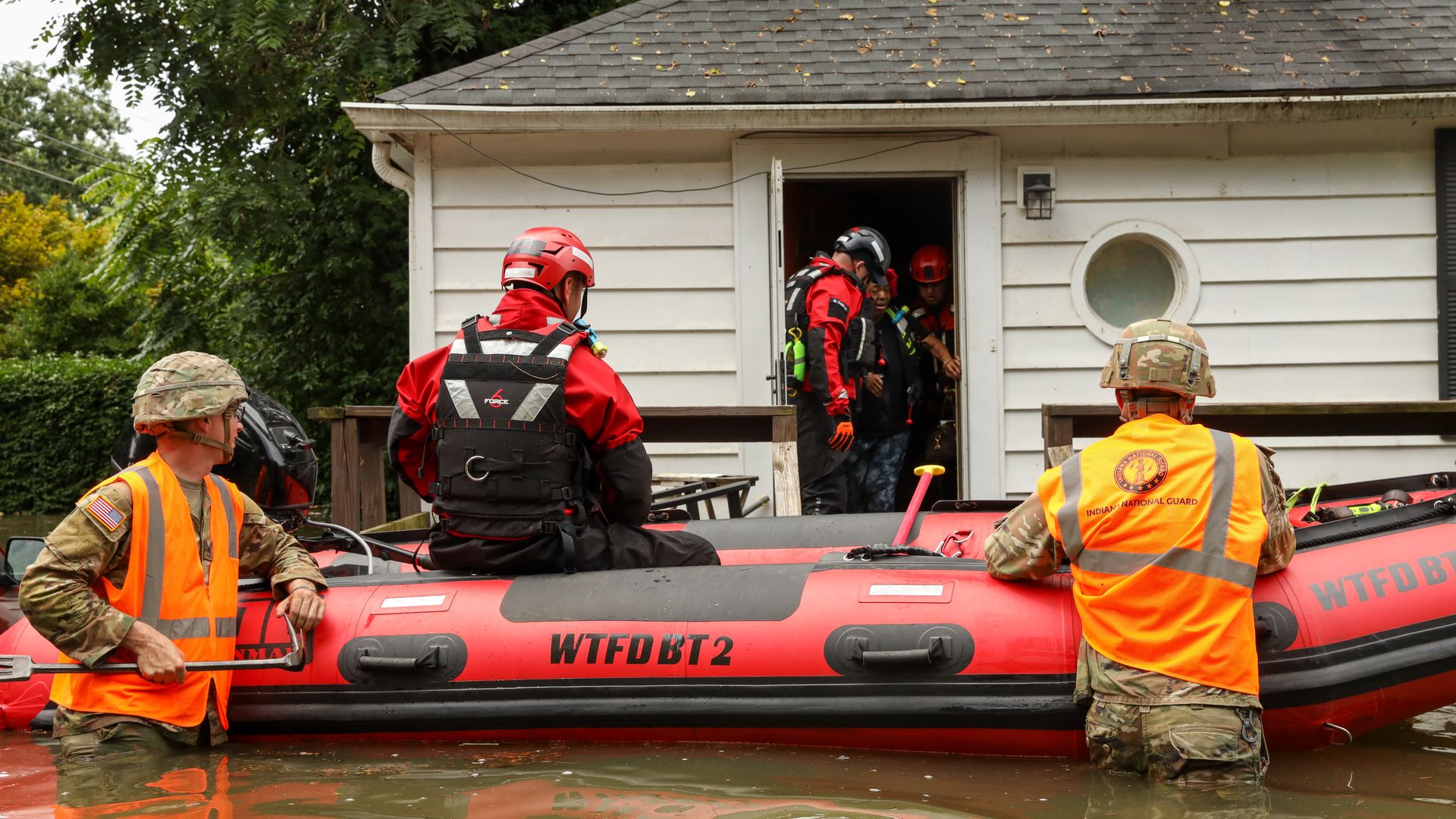 Rescue scene: orange-vested responders and helmets load into a red inflatable boat in a flooded yard; a white house with a gray roof and open door stands behind them.