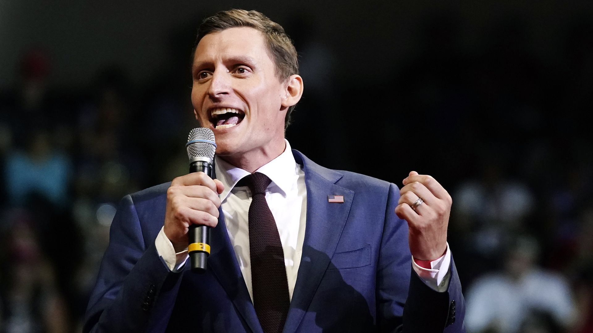 A man in a suit with an American Flag pin speaking into a microphone.