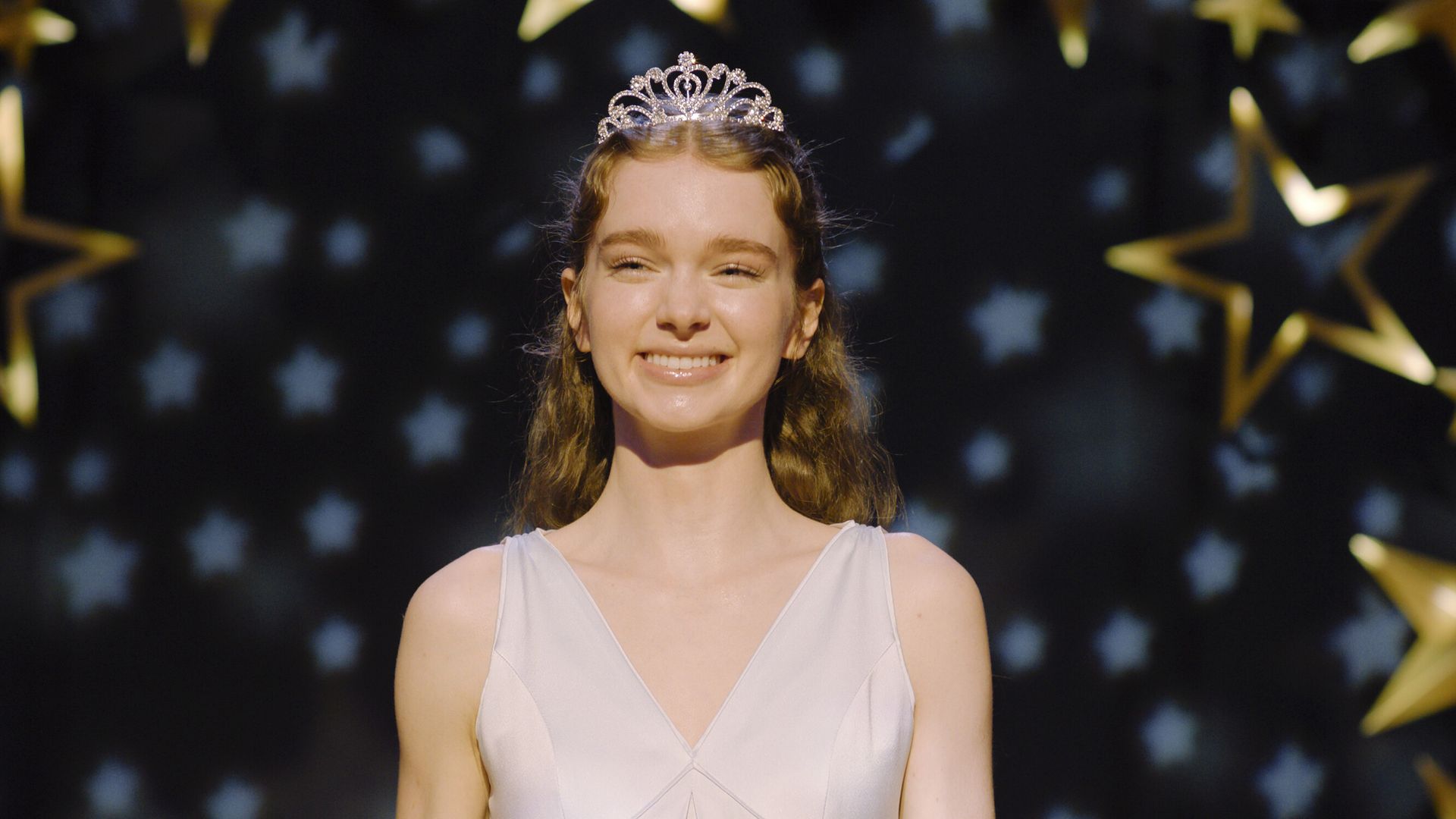 A young woman with red hair, spotlit and in a tiara, smiles.