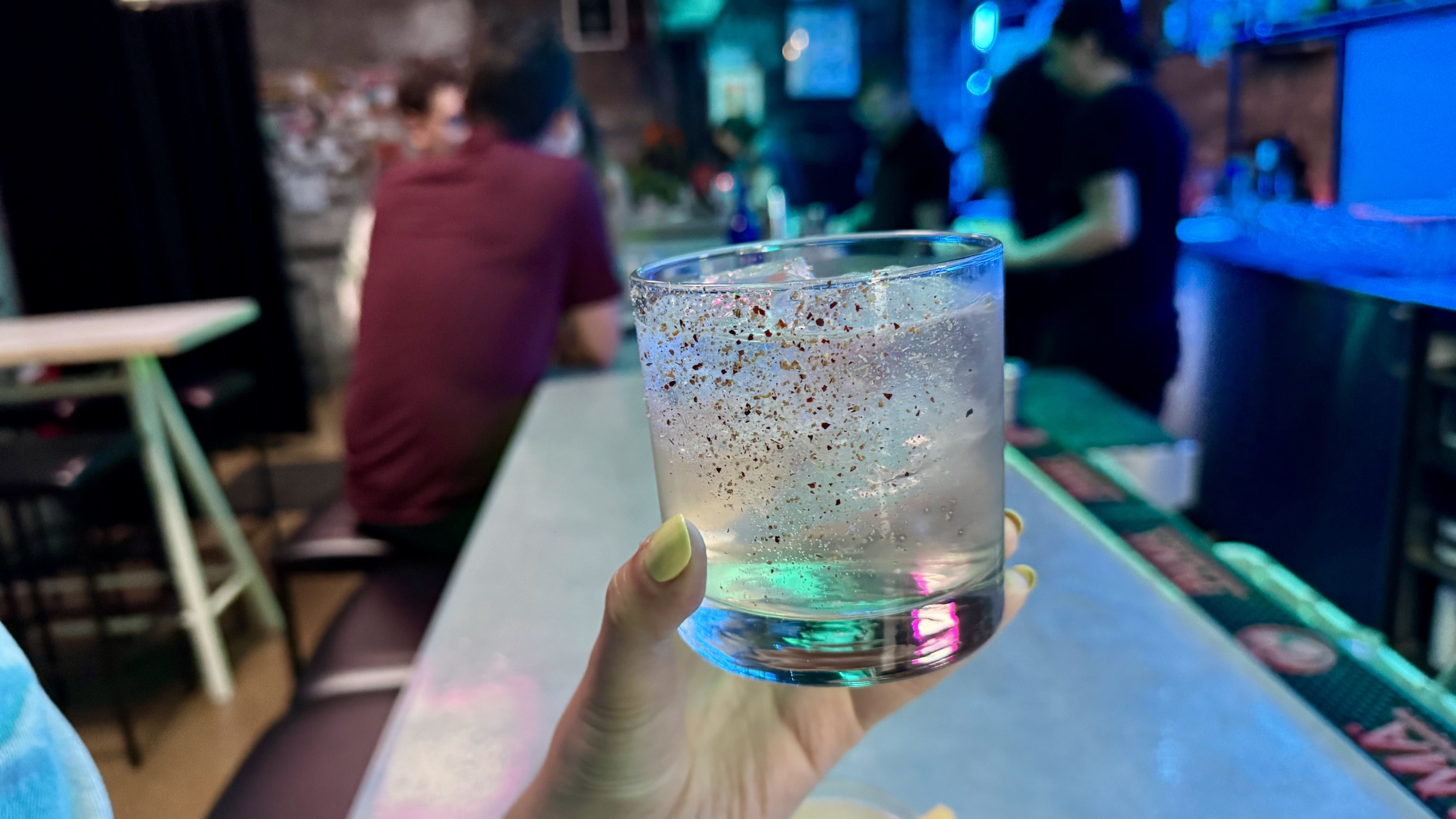 A clear drink with a tajin accent, being held against a background with neon lights