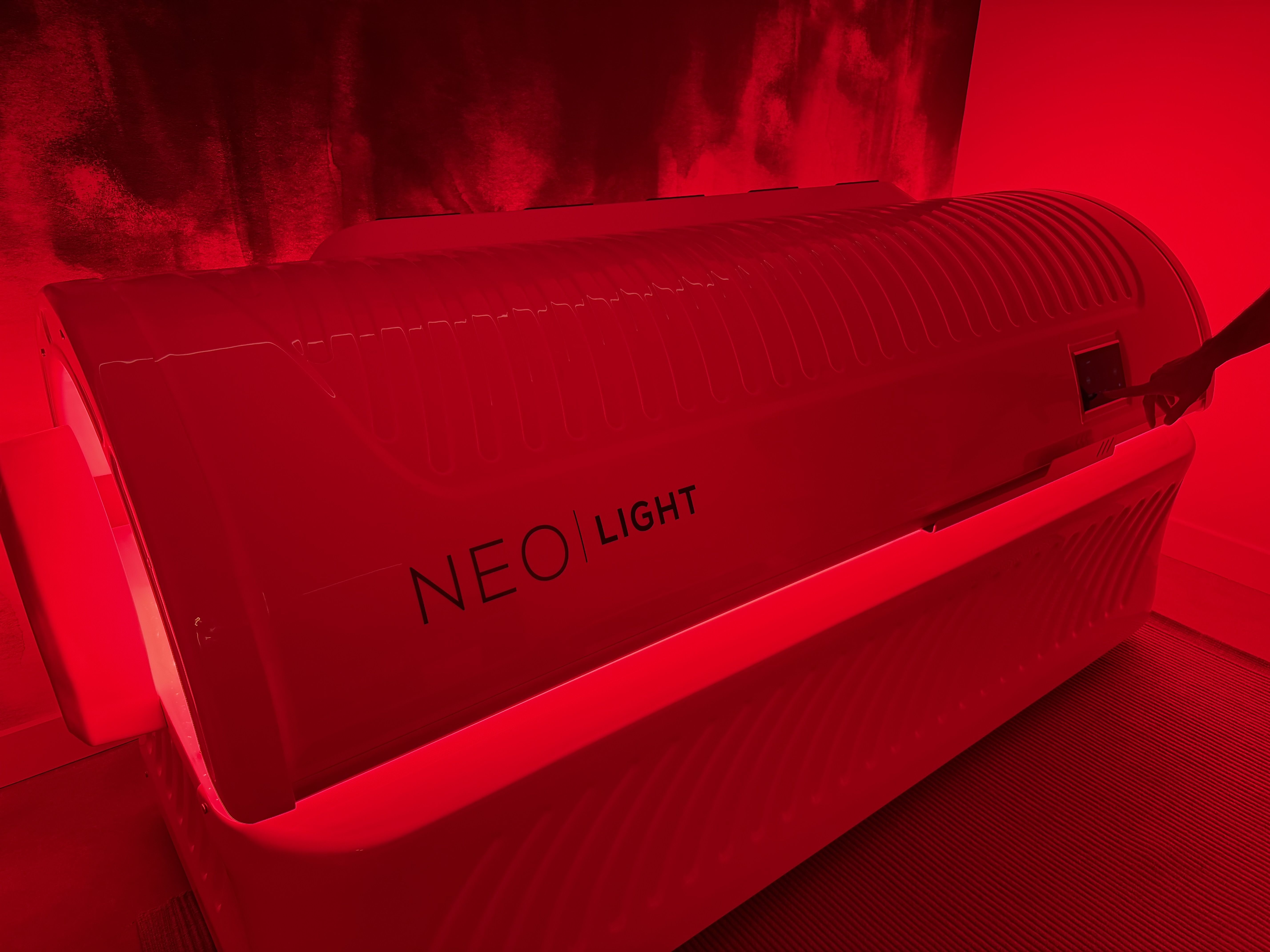 A red-lit NEO LIGHT therapy device with a person using a touchscreen on its side in a dimly lit room.