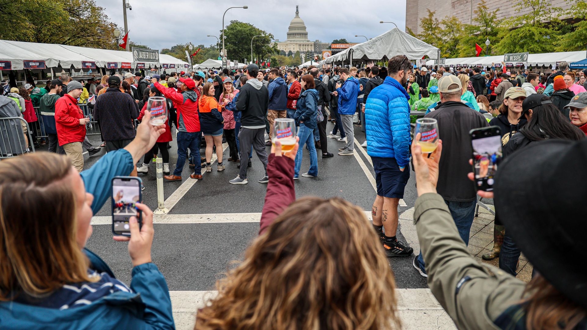 A photo showing the back of three people's heads raising glasses in the air. In front of them is a crowd of people and tents. It's overcast and most people are wearing coats. The U.S. Capitol building is visible in front of them.