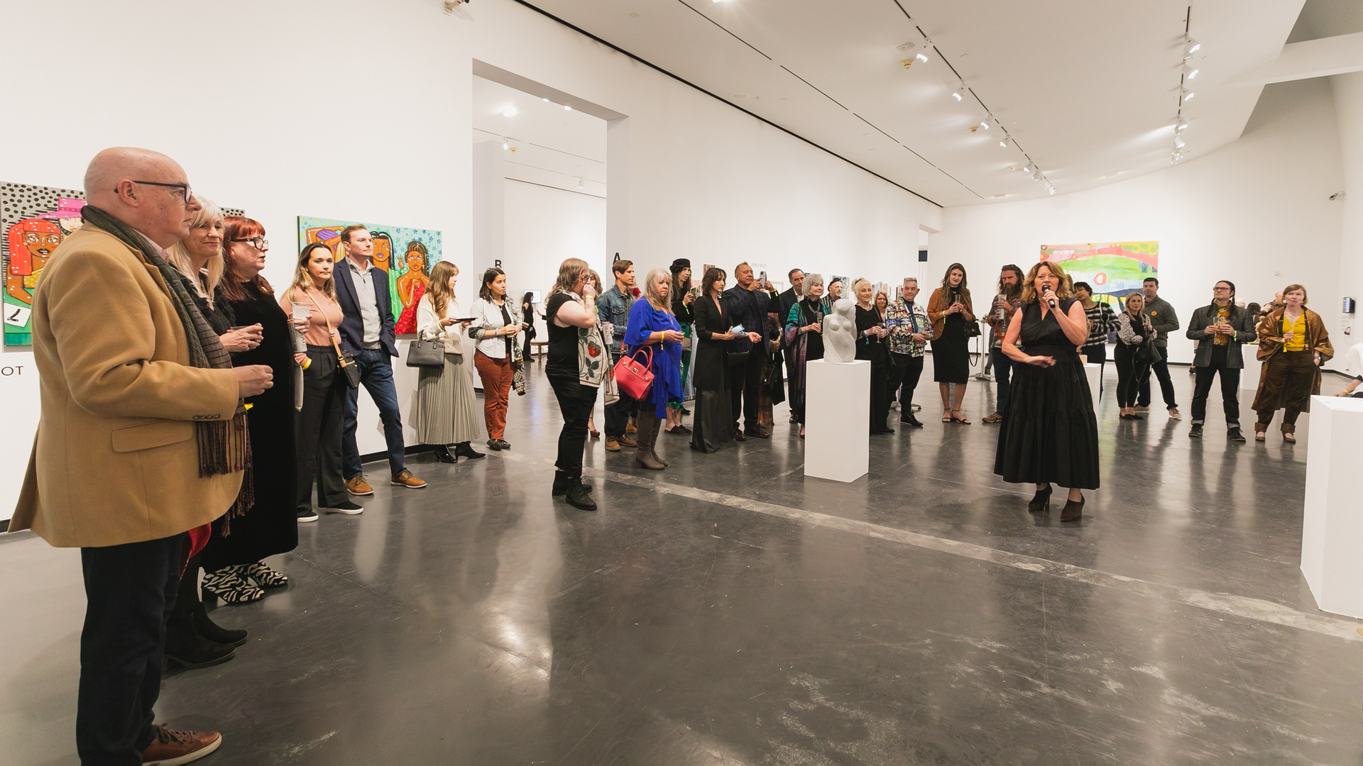 A woman in a black dress speaks to a group of a few dozen people gathered in a half circle around her in a white industrial space with art on the walls.