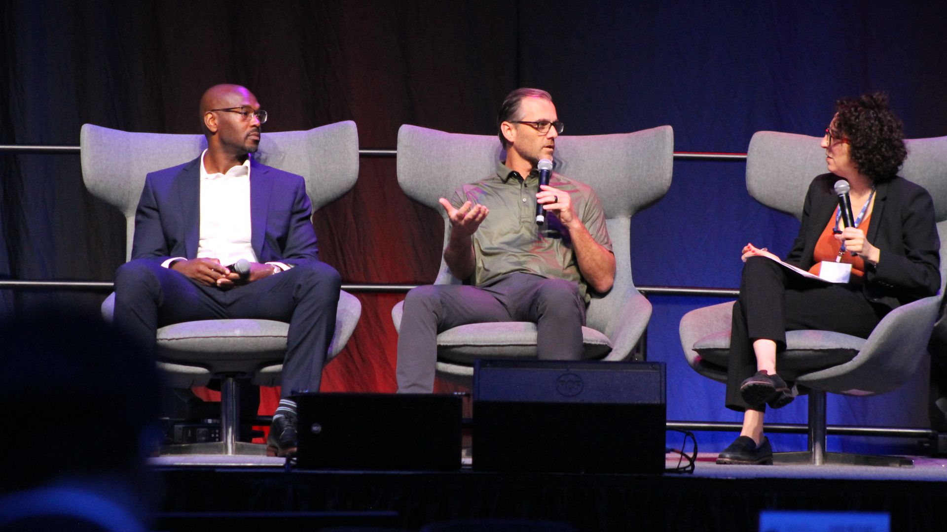 Three people seated on stage in gray chairs during a panel discussion, two men and one woman, all holding microphones, with dark curtains in the background under blue and red lighting.