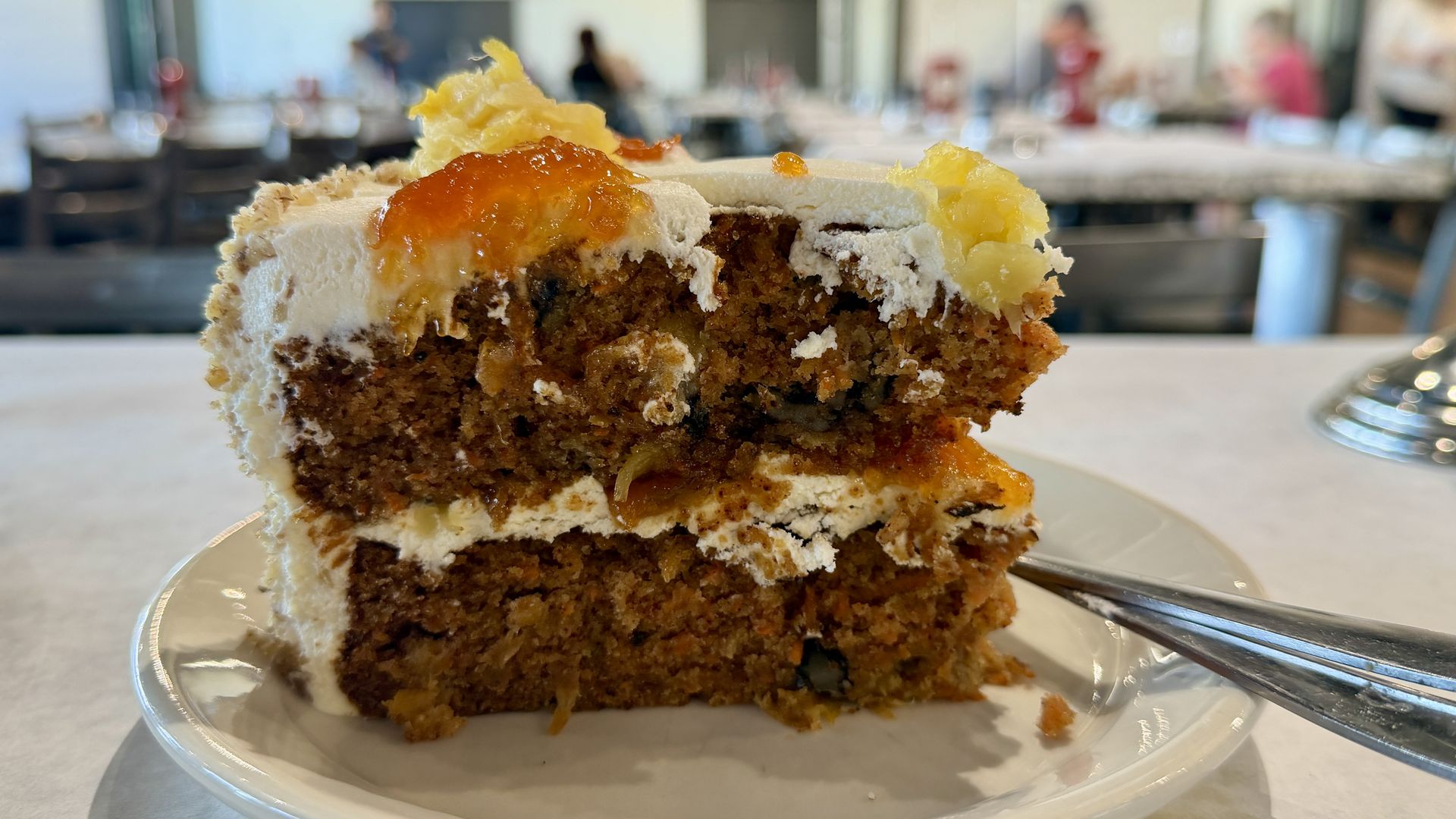 Close-up of a slice of carrot cake with white frosting topped with orange and yellow fruit preserves on a white plate with a fork, indoor restaurant setting blurred in the background.