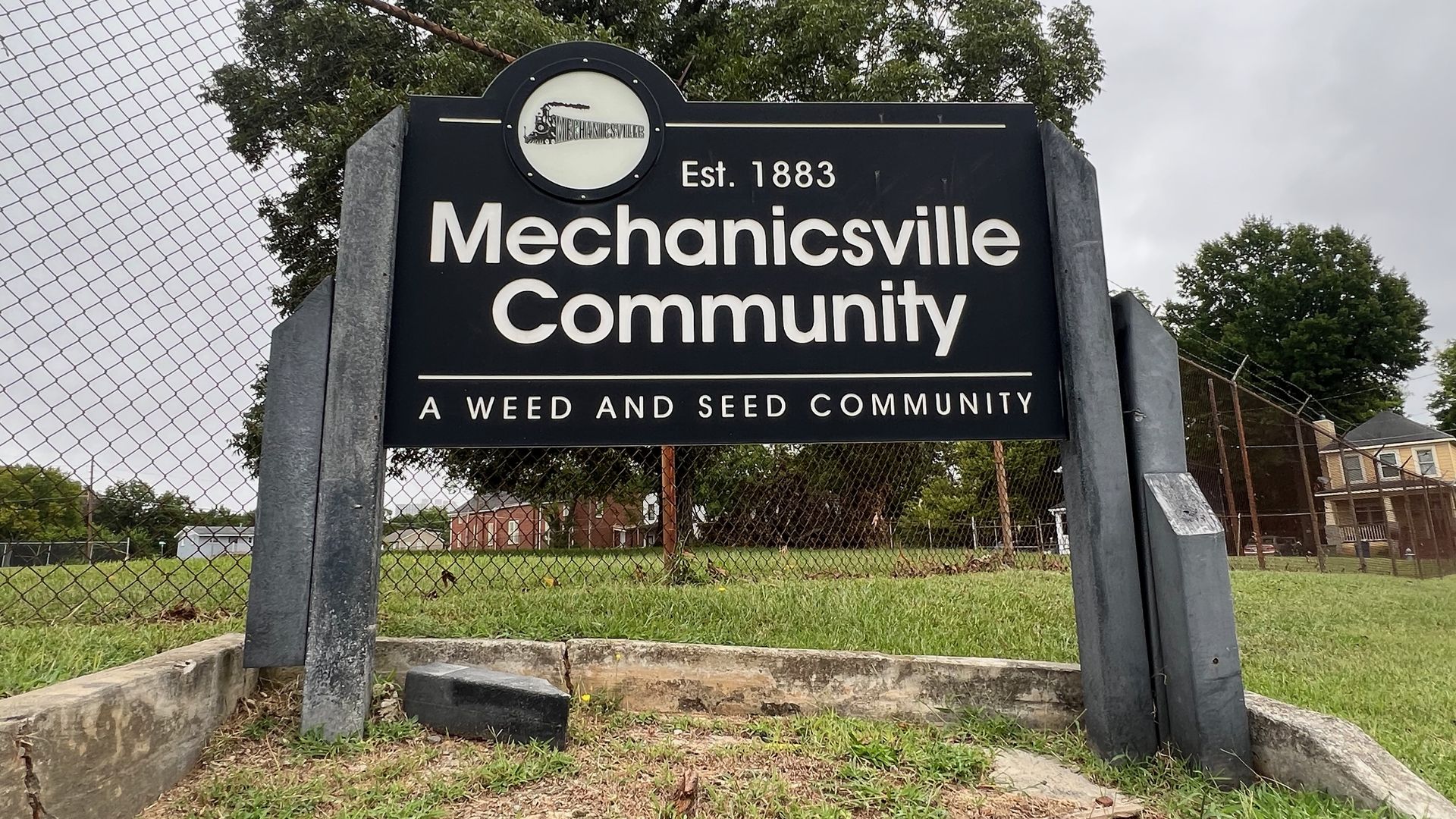 Sign reads Mechanicsville Community with Est. 1883 and A Weed and Seed Community; circular logo above, between metal posts, beside a chain-link fence on a grassy lot with trees in the background.