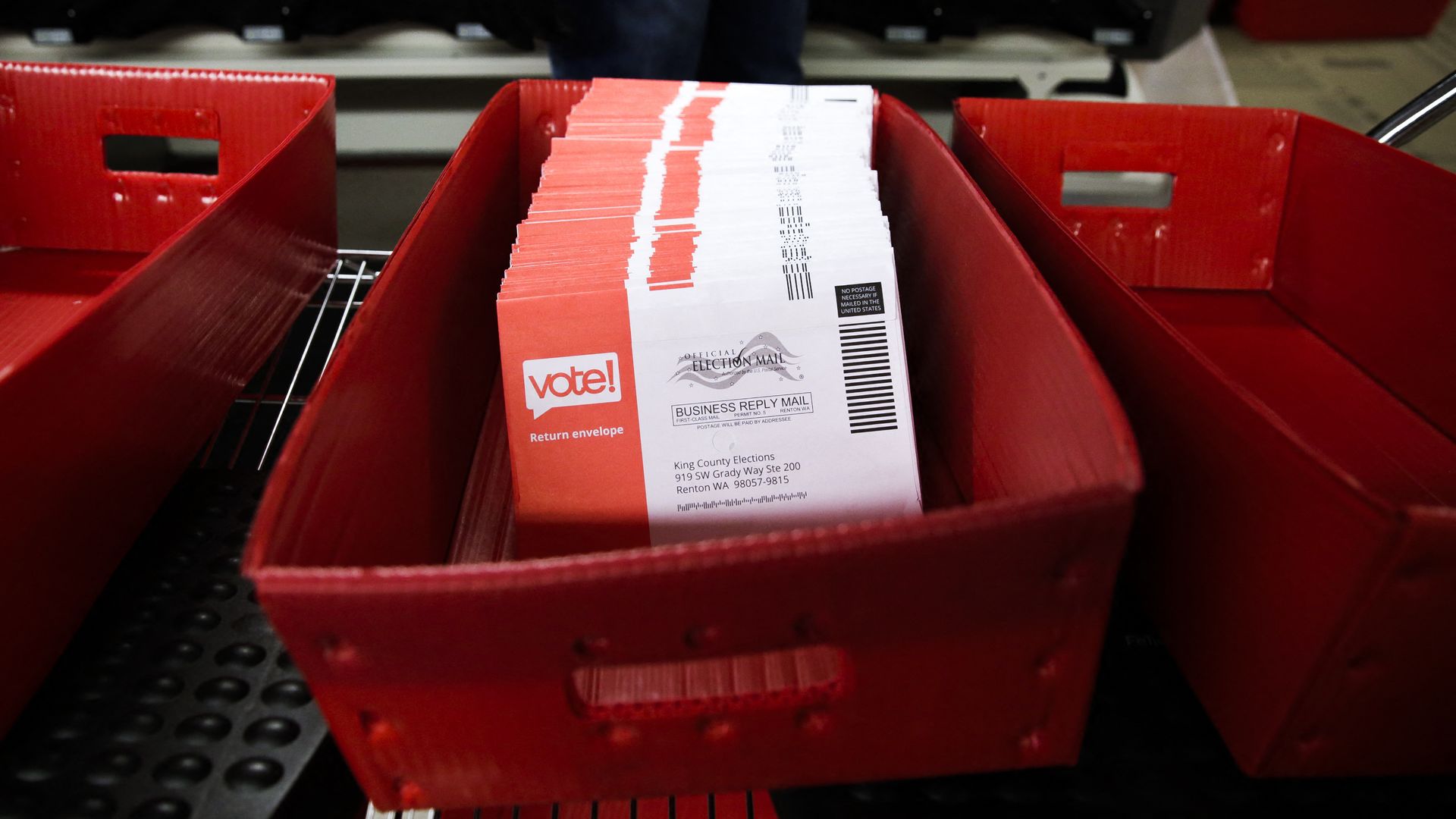 Red ballot drop boxes hold a stack of white and red "vote!" envelopes labeled "Official Election Mail" with barcodes, at a polling station.