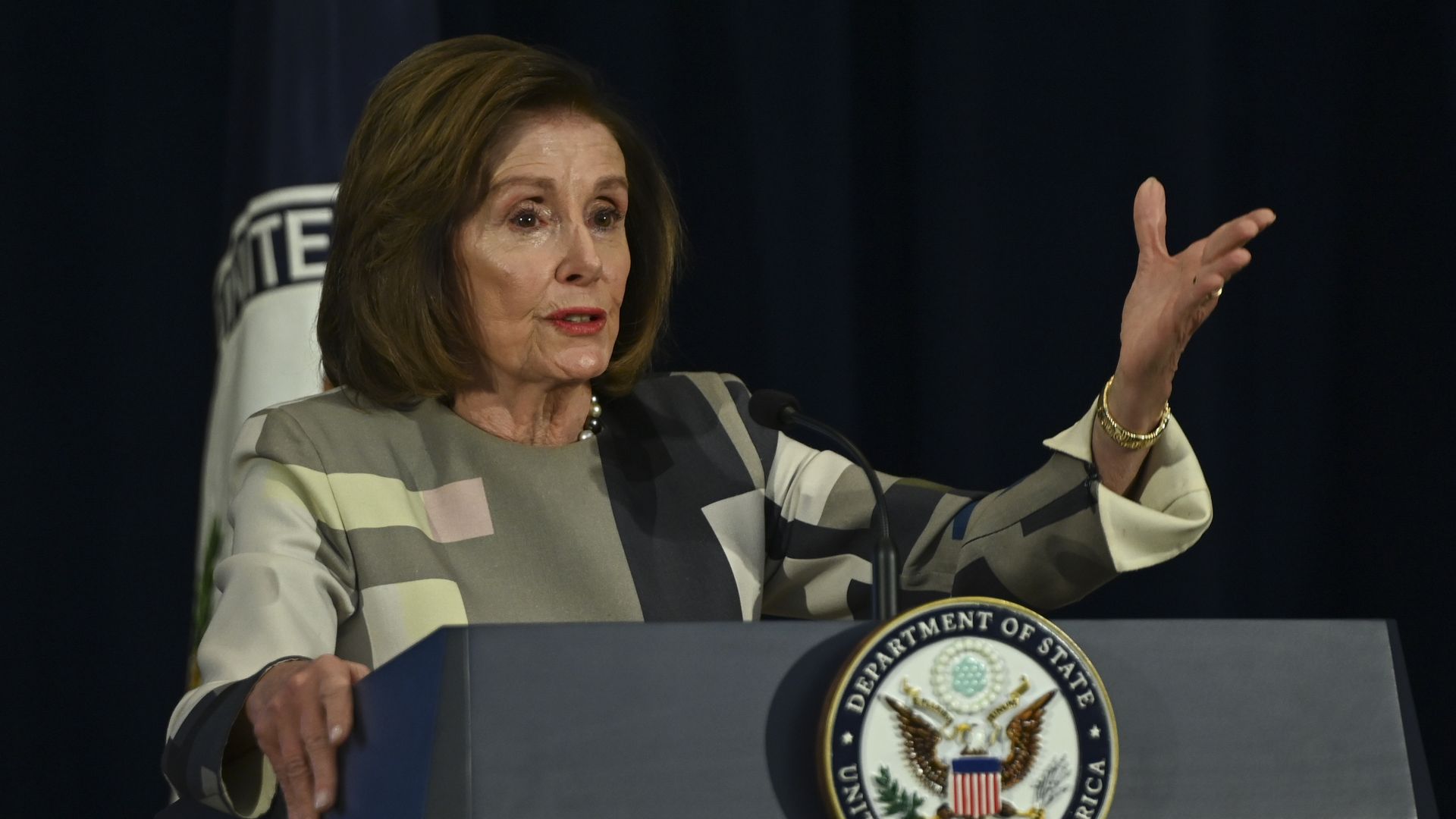 Nancy Pelosi wearing a gray dress and speaking at a podium with the state department seal.