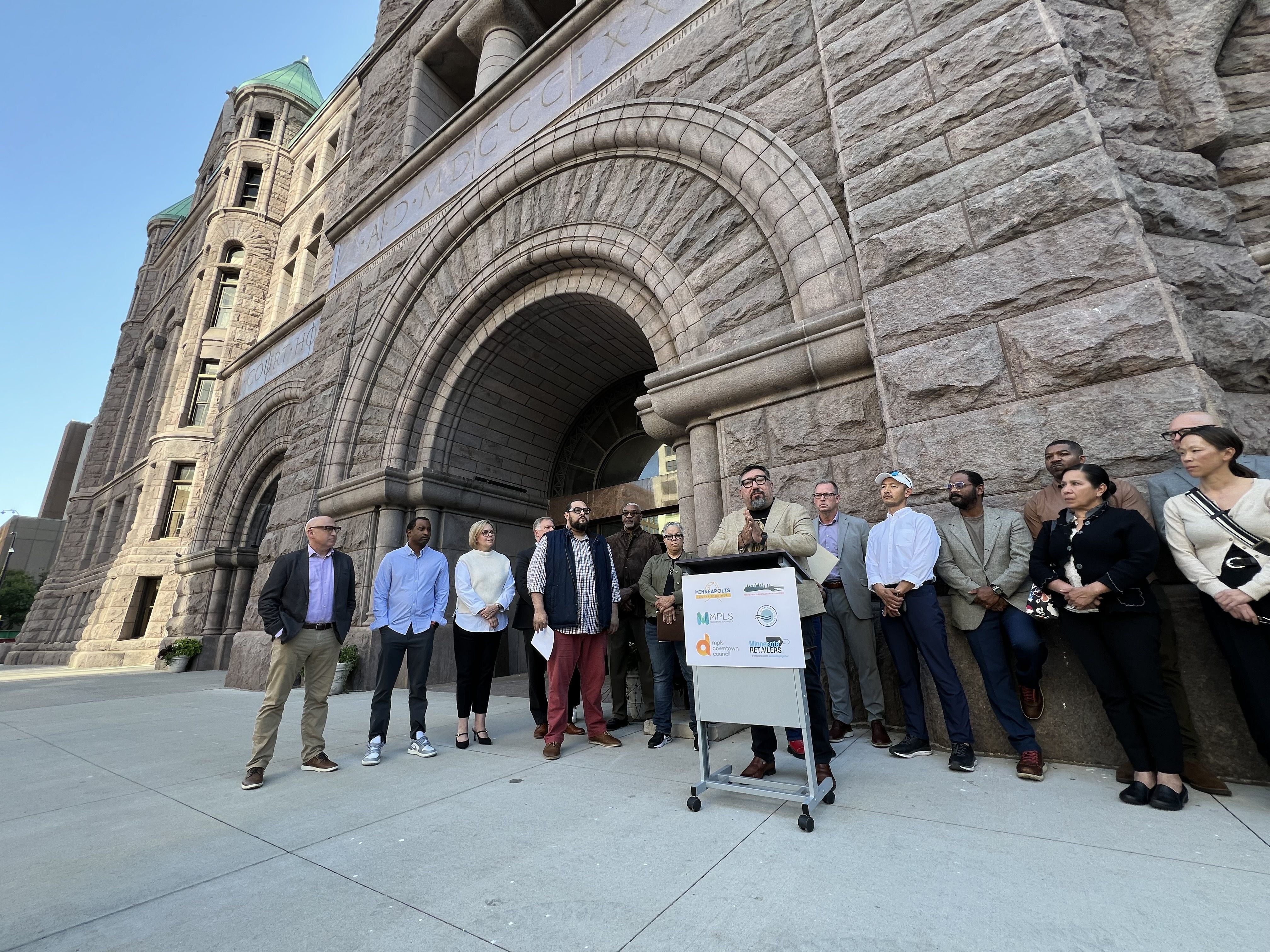 A group of people stand at a podium on a sidewalk outside a large stone building