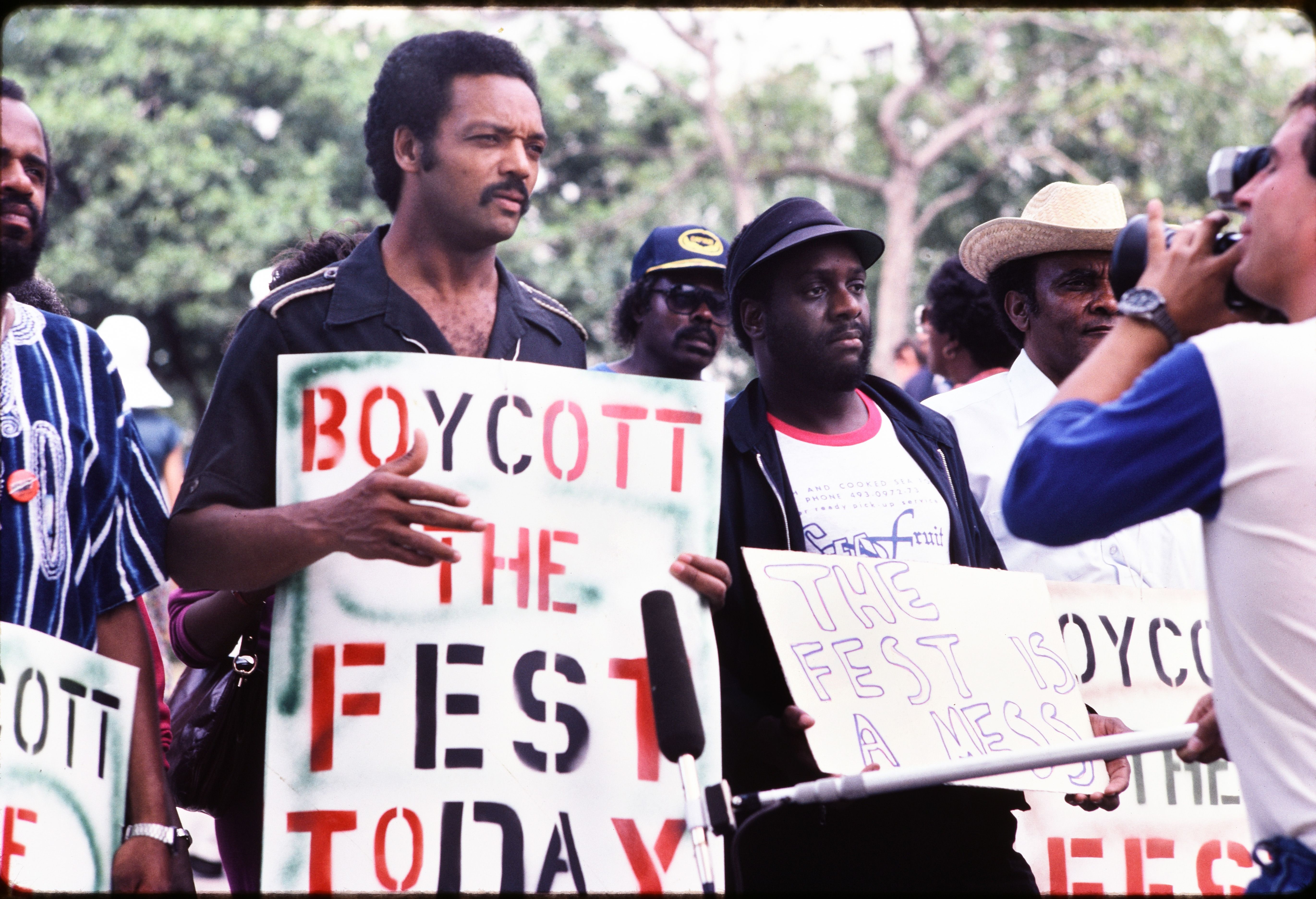 Group of African American men holding protest signs outdoors, one reads "BOYCOTT THE FEST TODAY" and another "THE FEST IS A MESS," while a man photographs them with a camera.