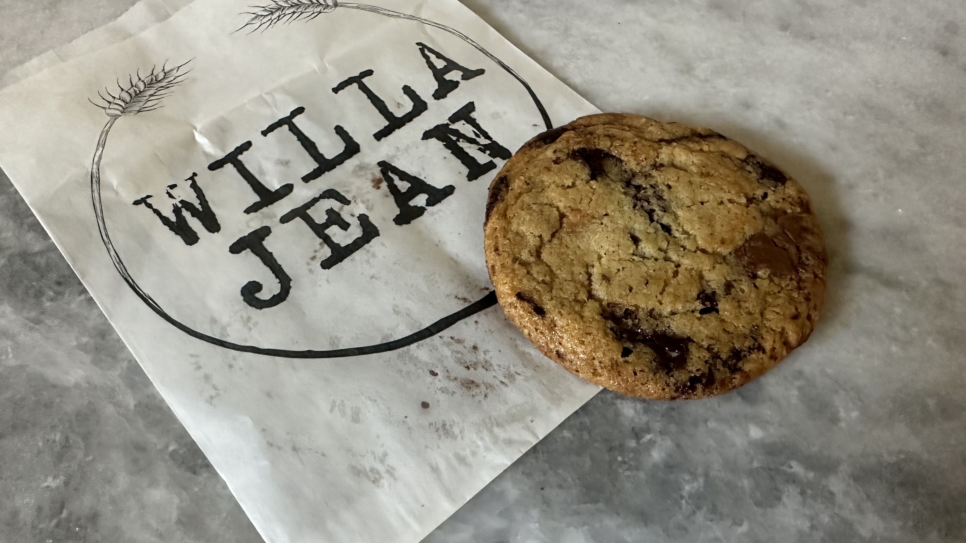 A chocolate chip cookies sits on a countertop next to a small pastry bag with the Willa Jean logo.