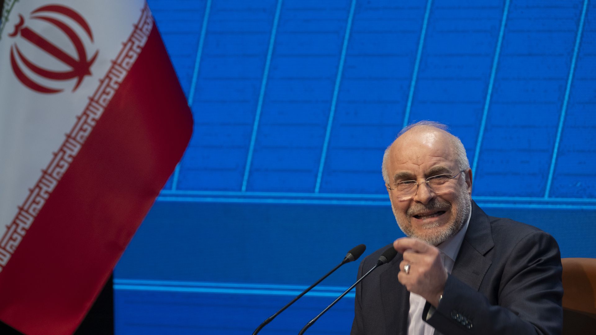Iranian Parliament Speaker, Mohammad Bagher Ghalibaf, a gray-bearded man in a dark suit speaks into two microphones at a conference; the Iranian flag is on the left and a blue geometric backdrop fills the background.