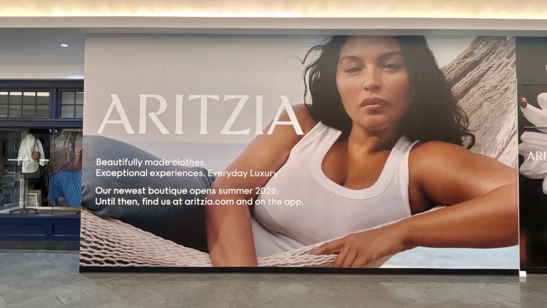 Advertisement for ARITZIA: a woman with dark hair in a white tank top lies on a wicker chair; large white "ARITZIA" text, with promotional copy about new boutique and online shop.