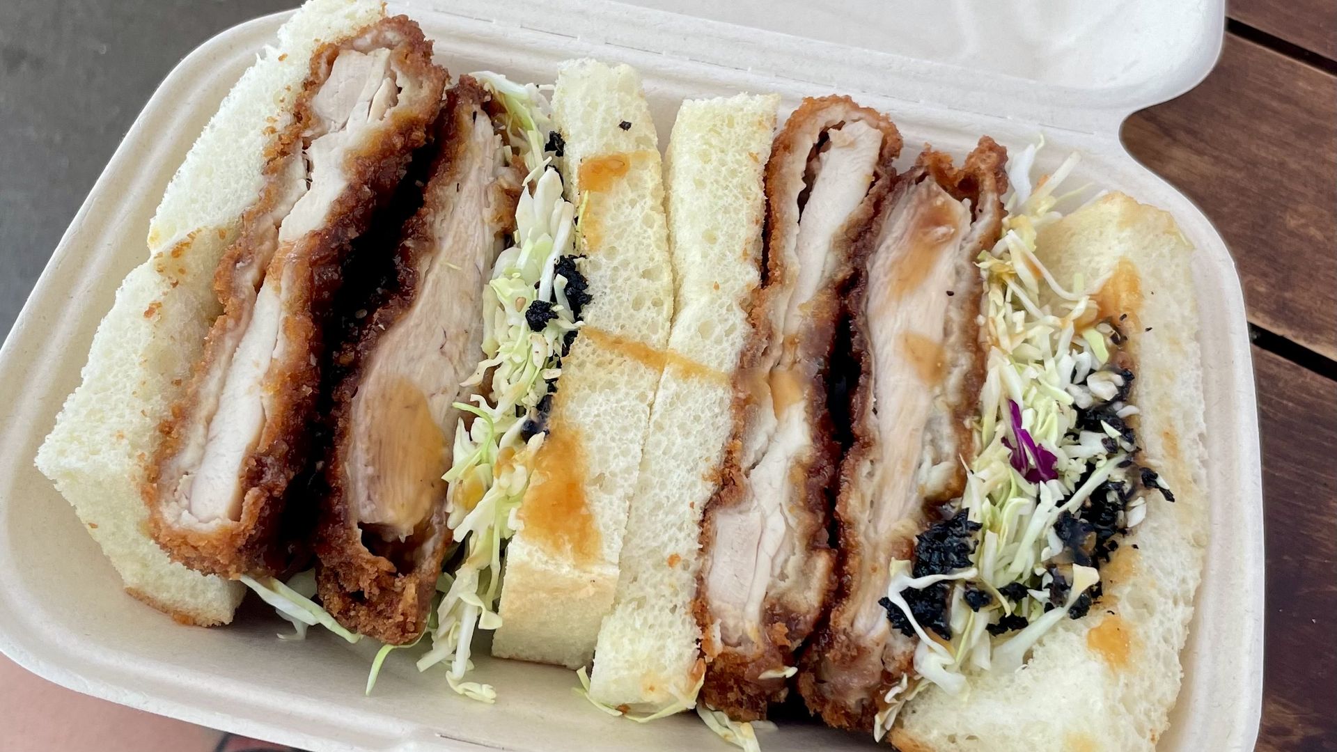 A takeout container with three thick-cut katsu sandwiches, filled with crispy breaded pork cutlet, shredded cabbage, dark sauce, and served between soft white bread slices.