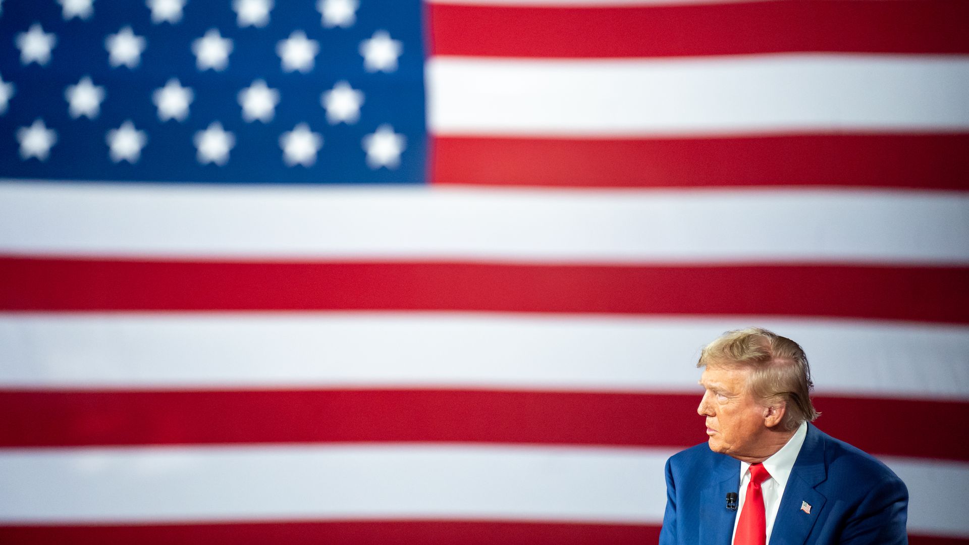 Donald Trump sits in front of a large backdrop of an American flag. He's wearing a blue suit, red tie and white shirt.