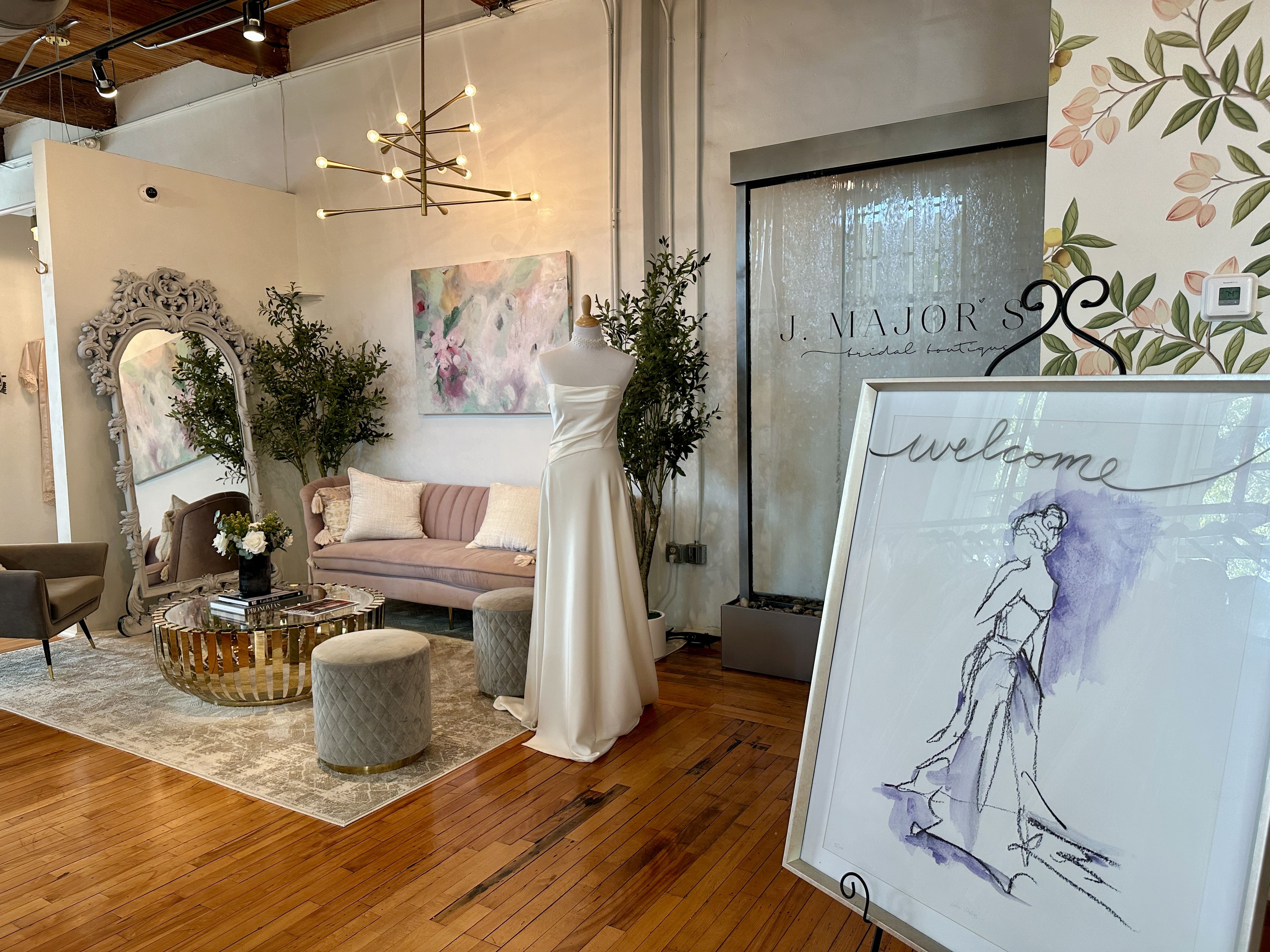 Elegant bridal boutique interior with a white strapless wedding gown on a mannequin, pastel pink velvet sofa, round gold coffee table, large ornate mirror, plants, and purple-accented welcome sign.