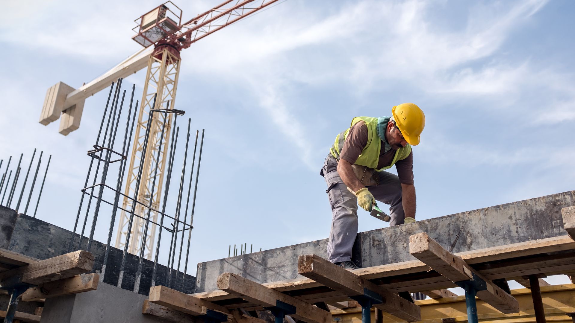 In this image, a construction working wearing a vest and hard hat works over a concrete beam. 