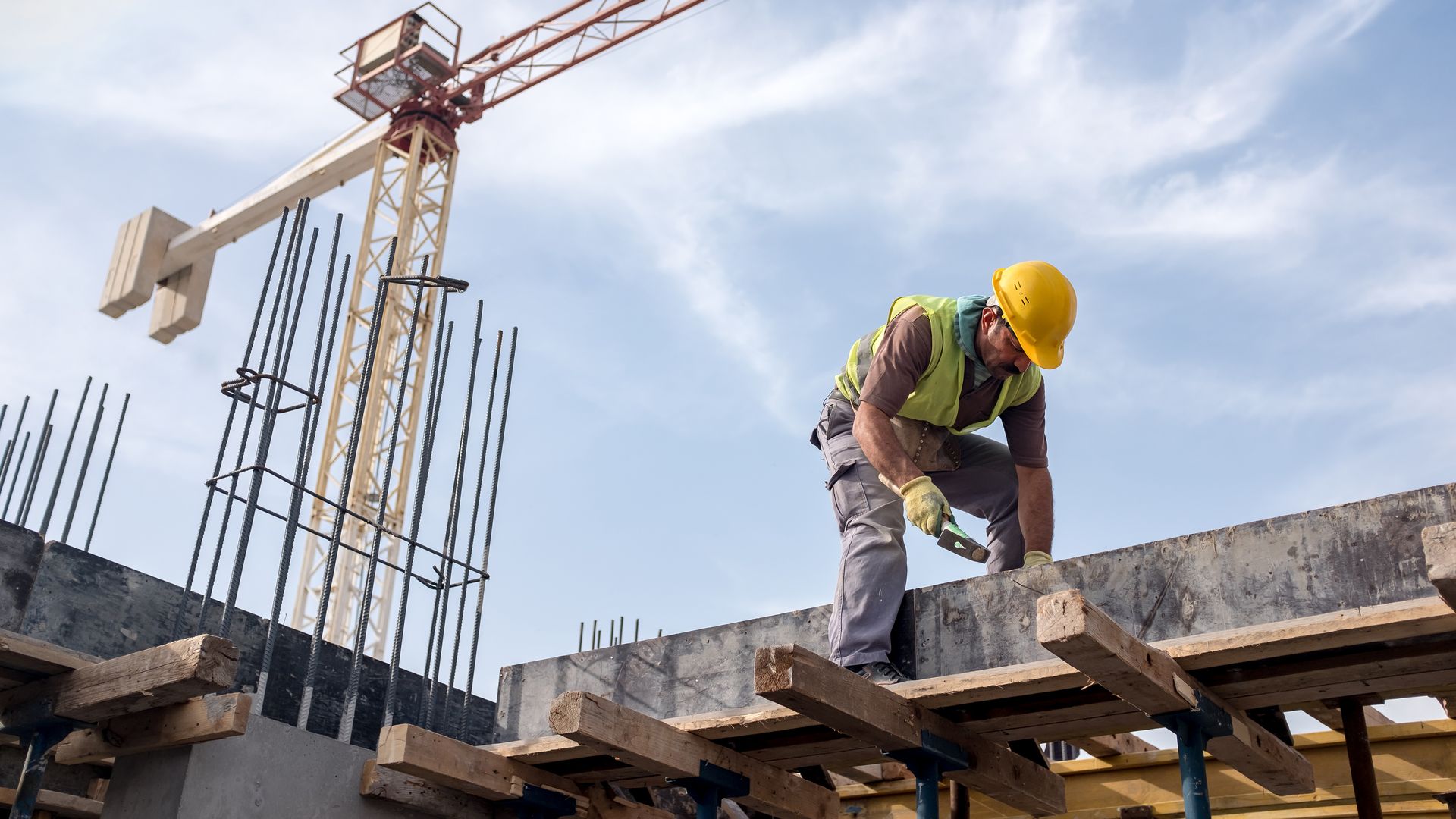In this image, a construction working wearing a vest and hard hat works over a concrete beam.