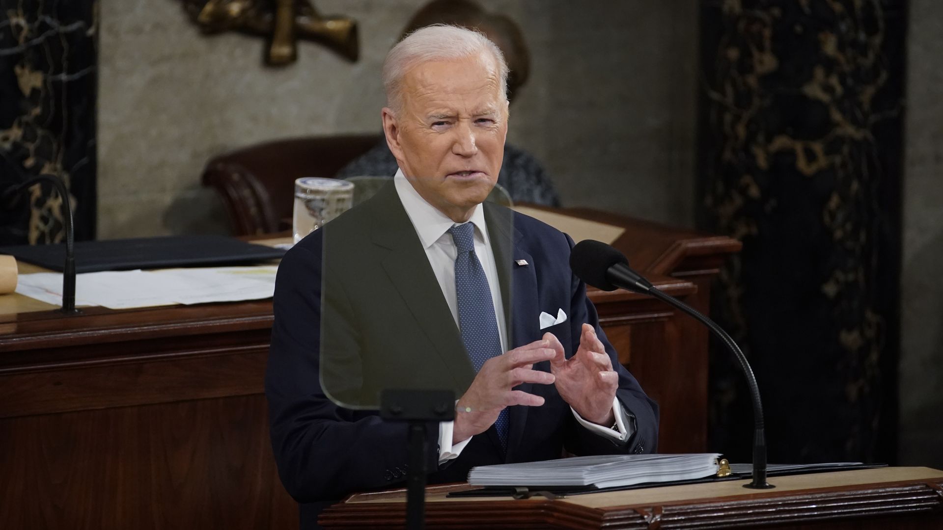President Biden, wearing a dark blue suit, white shirt and blue tie, speaks at the House of Representatives rostrum in front of a teleprompter.