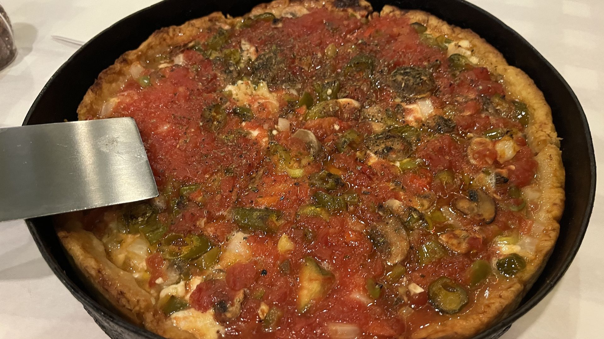 Thick-crust pizza in a dark round pan, topped with red tomato sauce, melted cheese, onions, and green peppers; a metal spatula rests along the pan's edge.