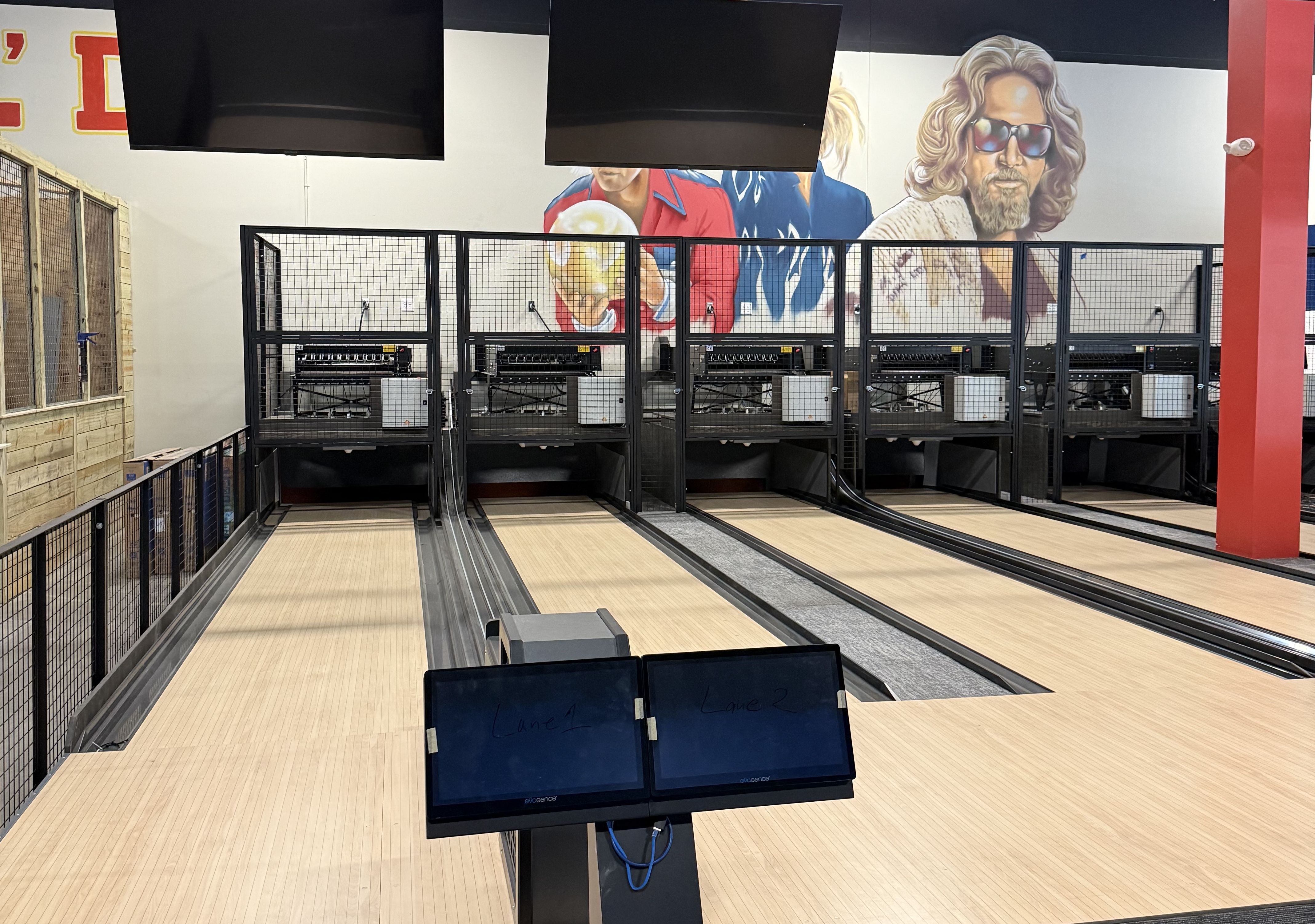 Empty bowling lanes in an indoor alley with wooden floors, black mesh cages for ball returns, large TVs above lanes, and a mural of a man with sunglasses on the wall.