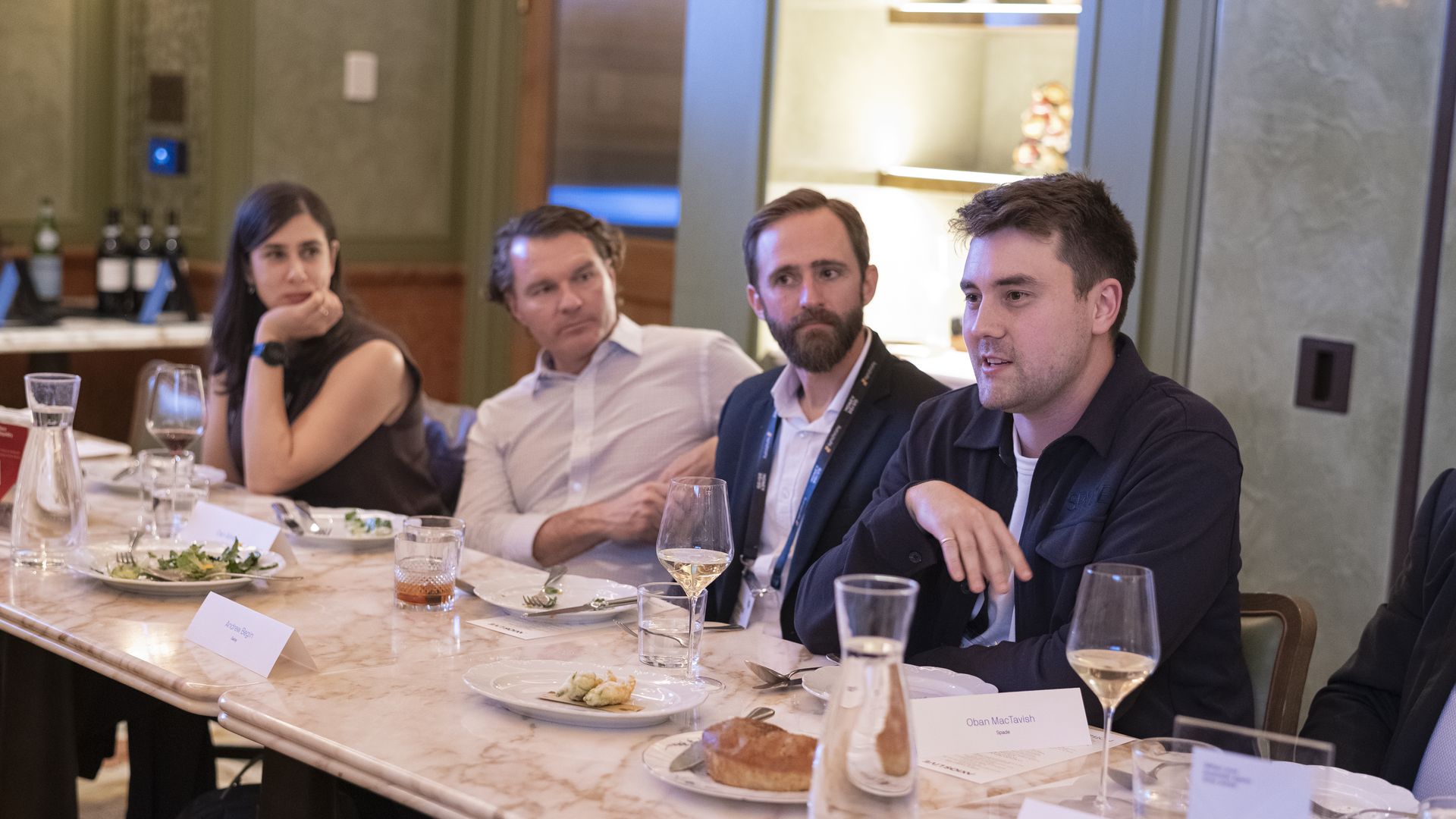 Four people sitting at a marble table with plates, glasses of wine and water, engaged in conversation in a softly lit dining room.