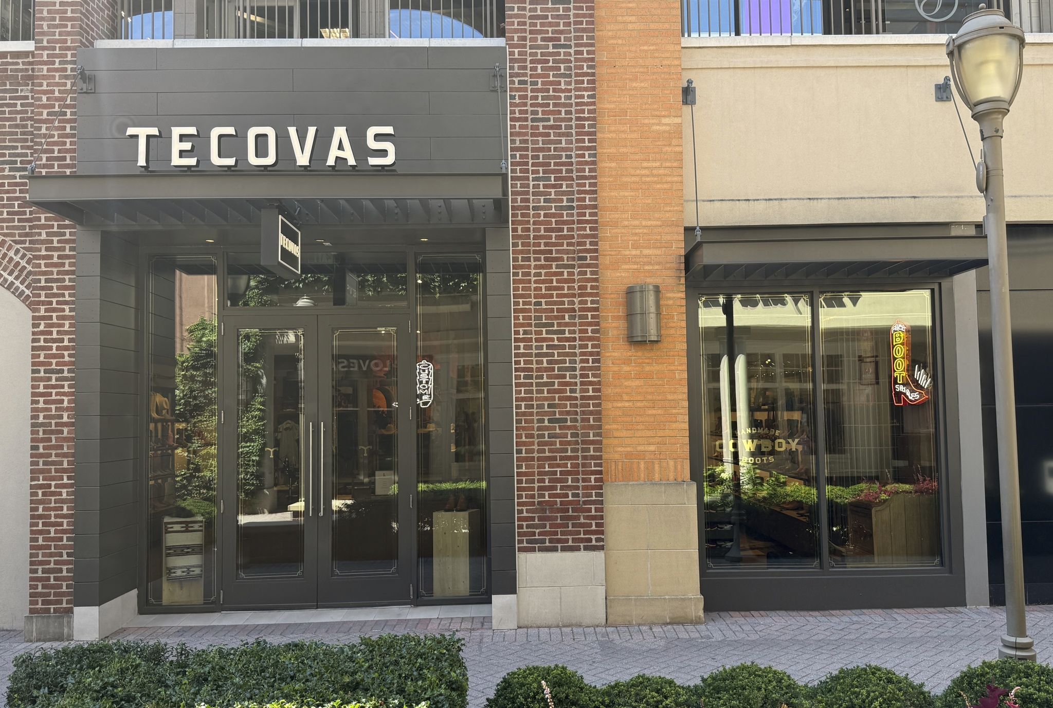 Storefront with a large gray sign that reads "TECOVAS" on a brick-gray facade; glass doors, a streetlamp on the right, neon "BOOTS" sign in the window, and greenery along the sidewalk.