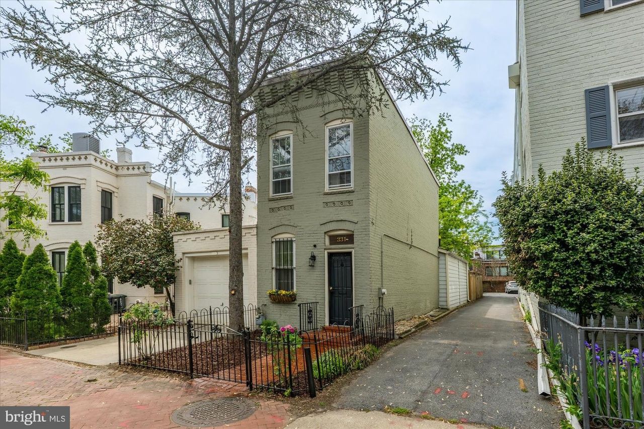 Two-story pale gray-green brick townhouse with a black front door and a small fenced garden. A tall tree stands in front; a driveway runs along the right side beside other brick homes.