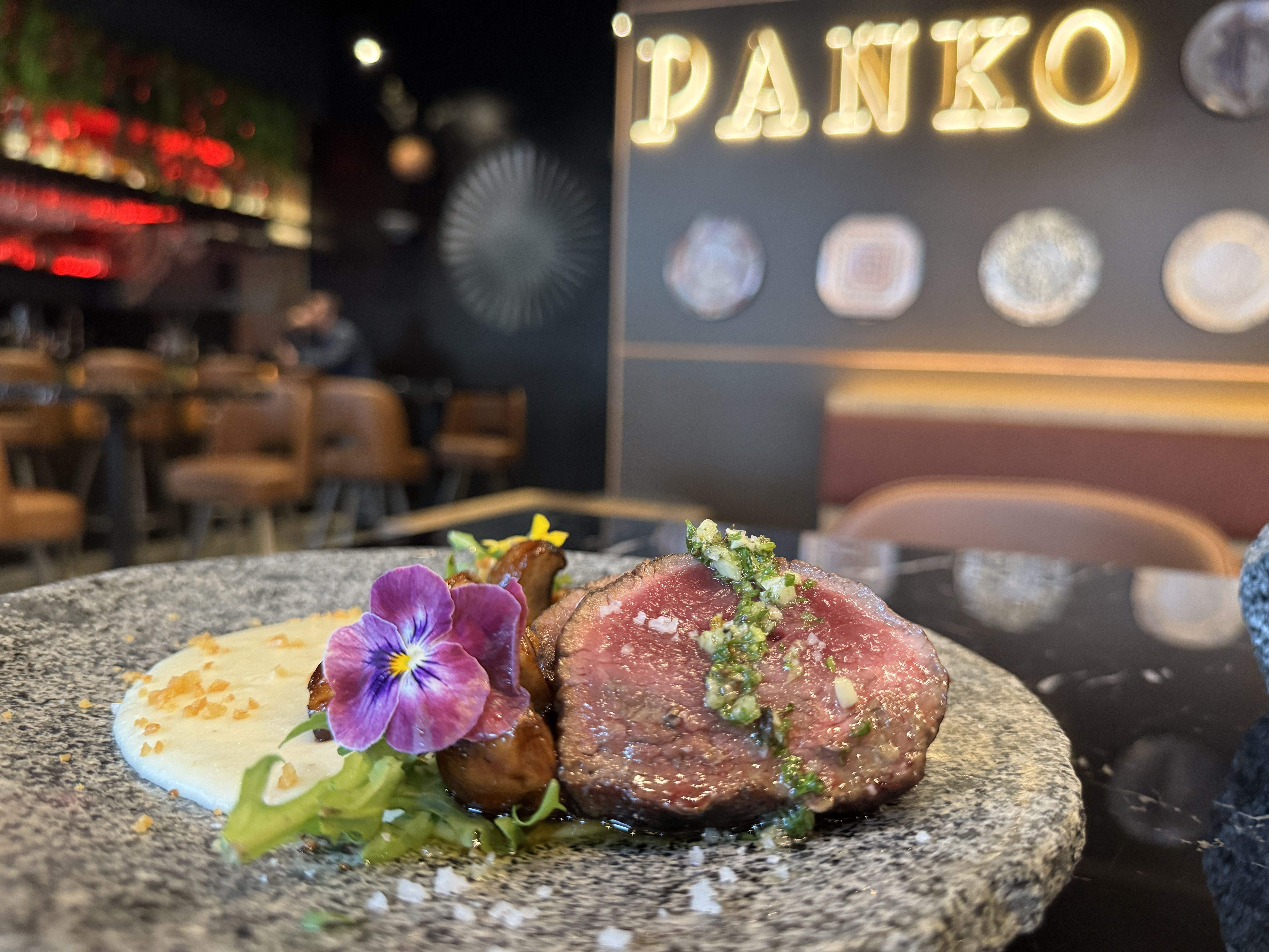 Close-up of plated gourmet dish with sliced medium-rare beef topped with green herb sauce, creamy white puree, sautéed mushrooms, greens, and a purple edible flower on a speckled plate, in a restaurant.