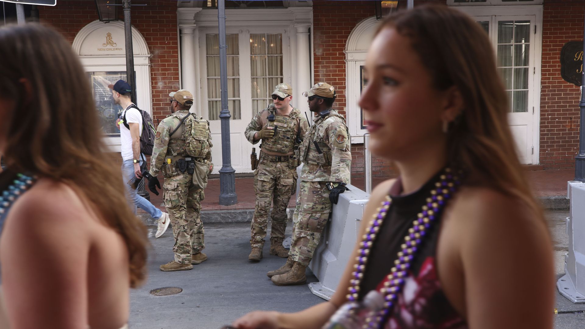 Two women in the foreground walk past the camera with Mardi Gras beads around their necks. In focus in the background, three National Guard soldiers stand.