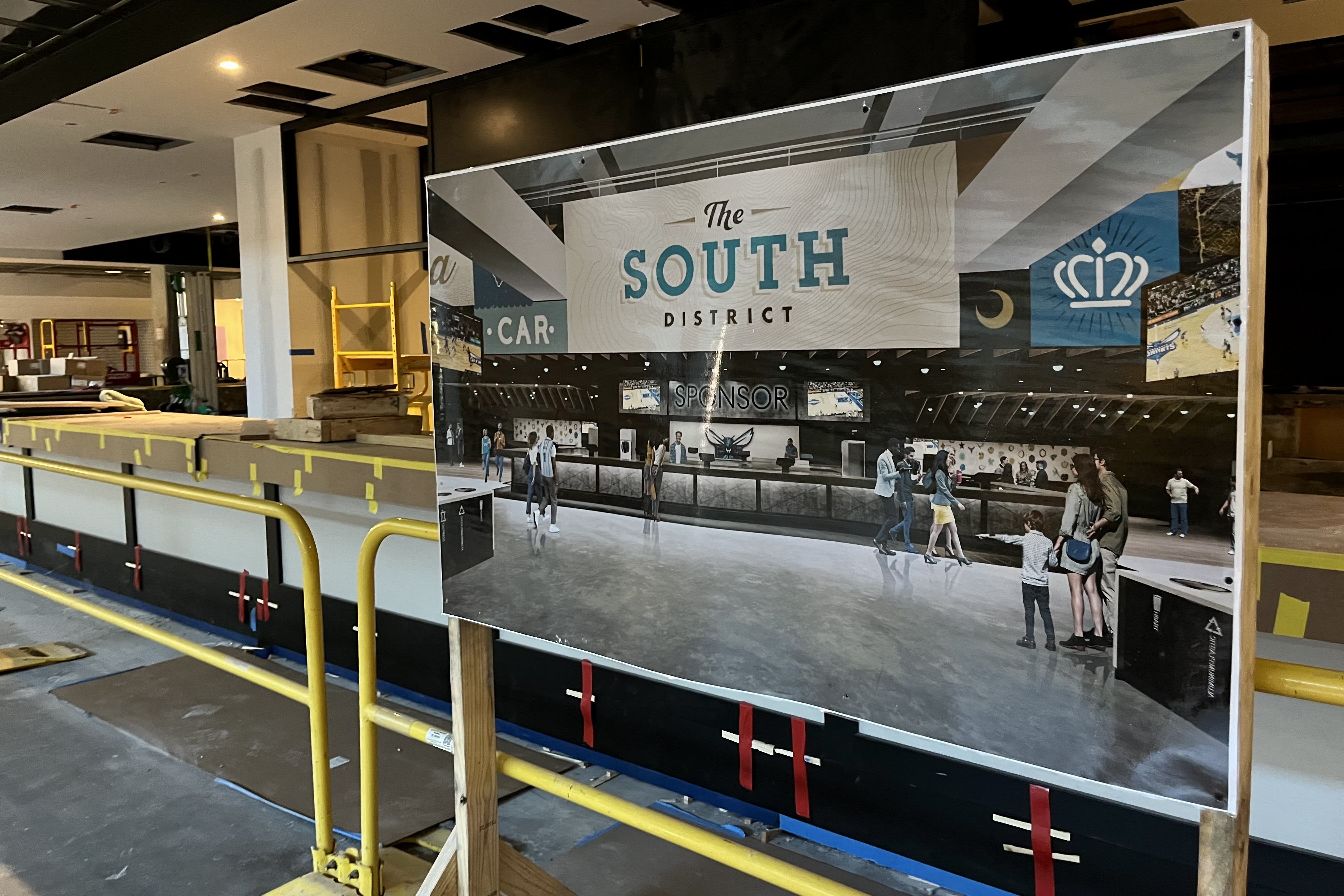 Construction site with a large sign showing a rendering of "The South District" lobby area featuring people walking and a reception desk under artificial lighting.
