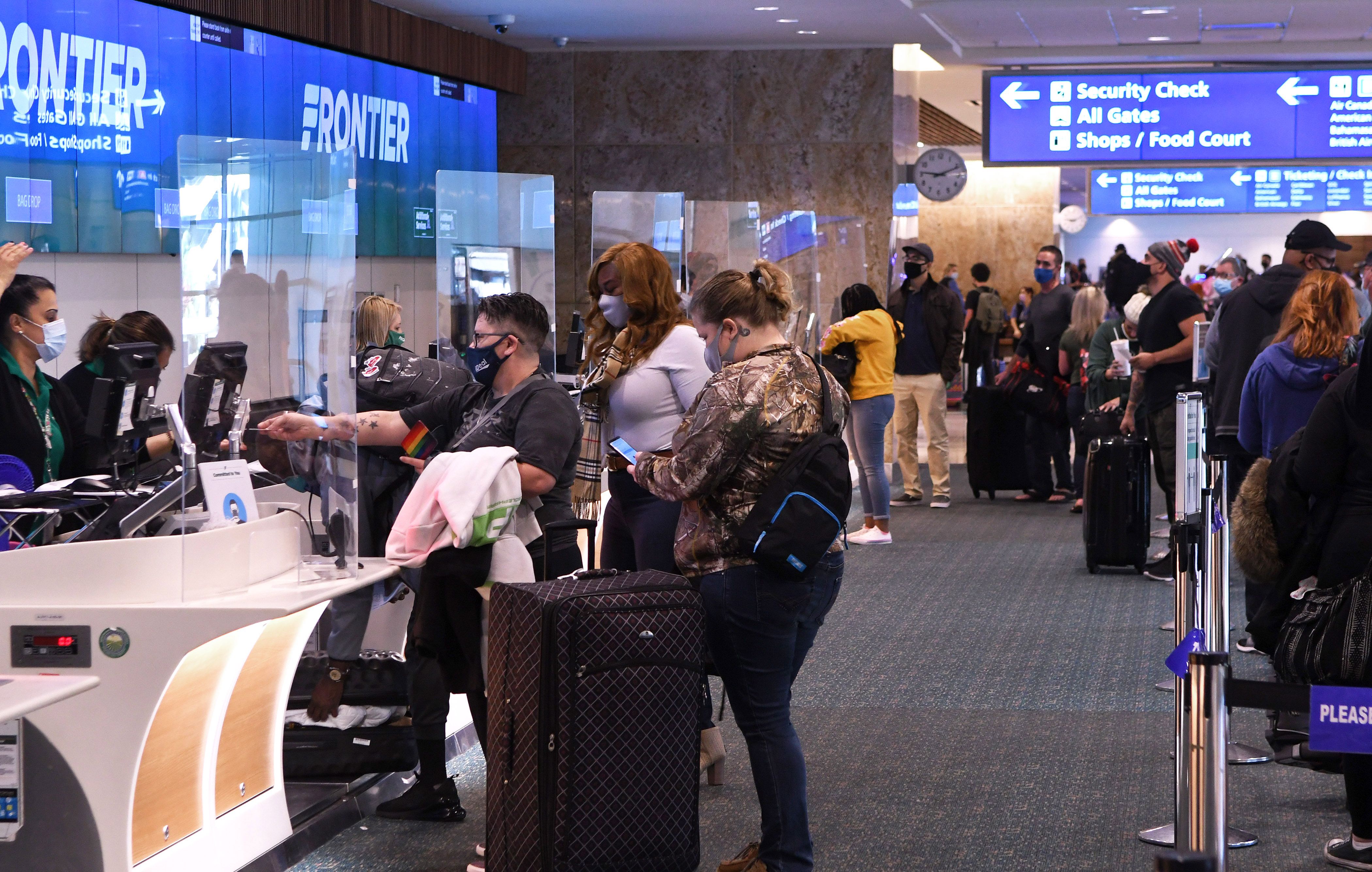 A crowd of people wearing masks and carrying mostly rolling suitcases stand at a check in counter with a Frontier sign 