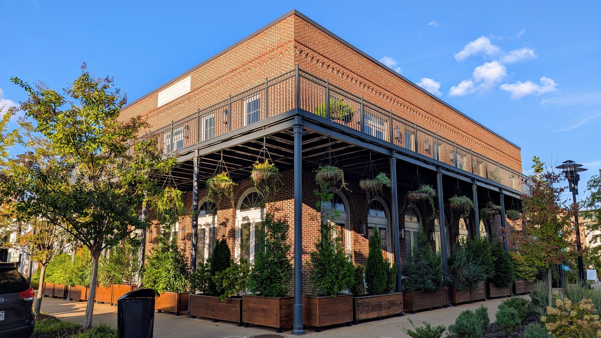 Two-story brick building with black iron balcony and hanging plants, surrounded by greenery and a clear blue sky, with a black trash bin and parked car nearby.