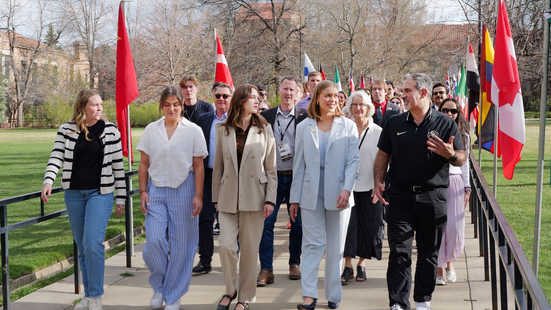A diverse group walks along a flag-lined path in a park, wearing business-casual outfits. Front-row women in light-colored suits smile as others follow.