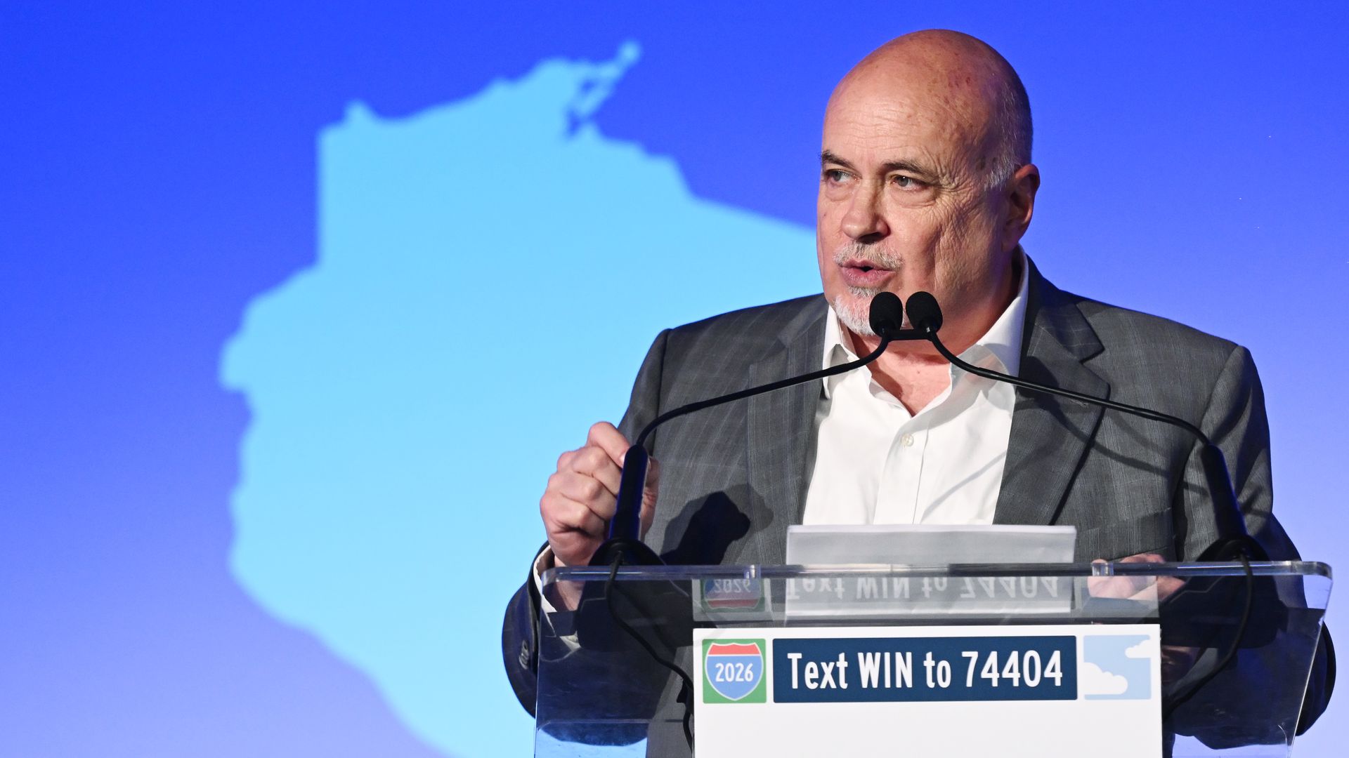 Rep. Mark Pocan, wearing a gray suit and white shirt without a tie, speaks at a podium in front of a blue outline of Wisconsin.