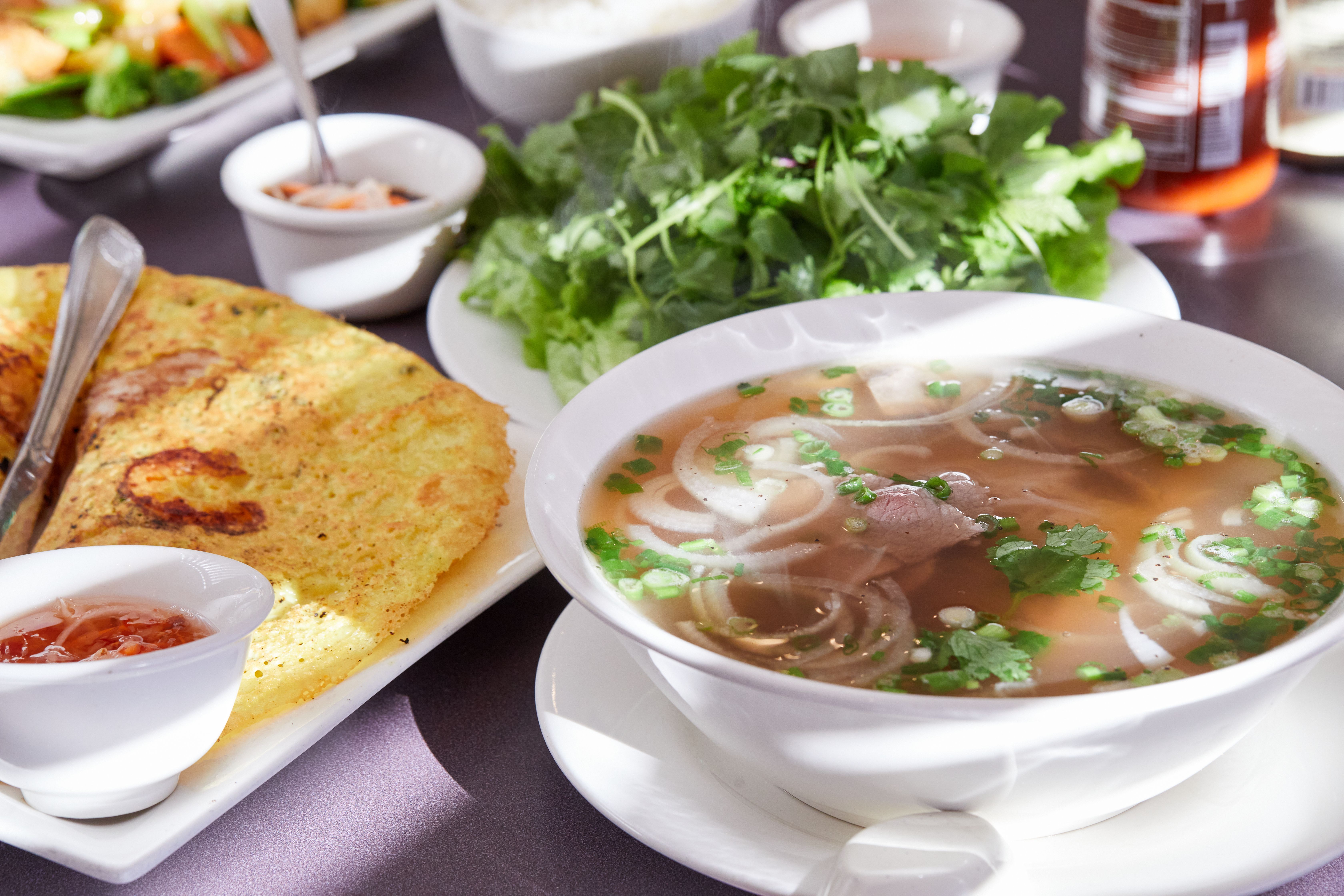 Vietnamese meal: clear-broth soup with beef and onions, a crispy omelet on a plate, a small cup of sauce and a bunch of cilantro and greens; condiments blurred in the background.
