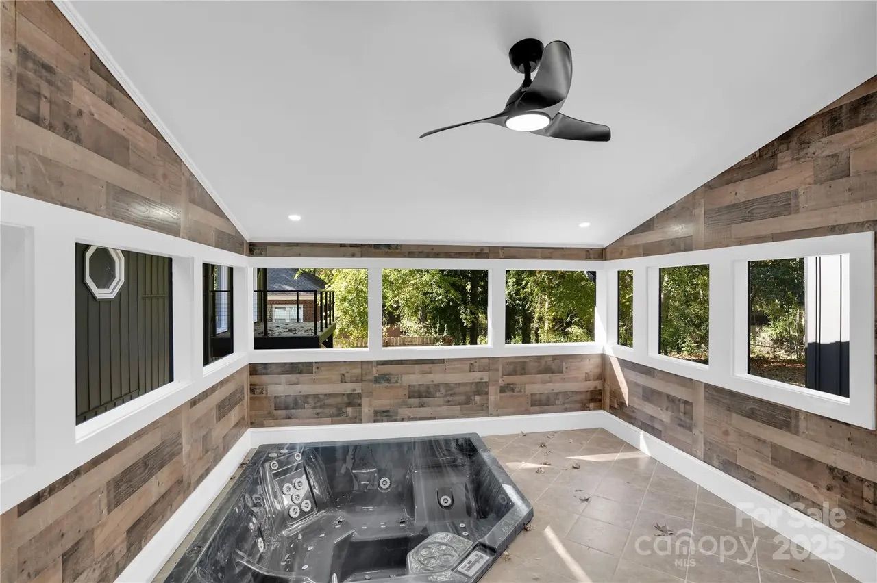 Enclosed patio with wood panel walls, high white ceiling with black ceiling fan, square tiled floor, and black hot tub beneath large windows showing outdoor greenery.