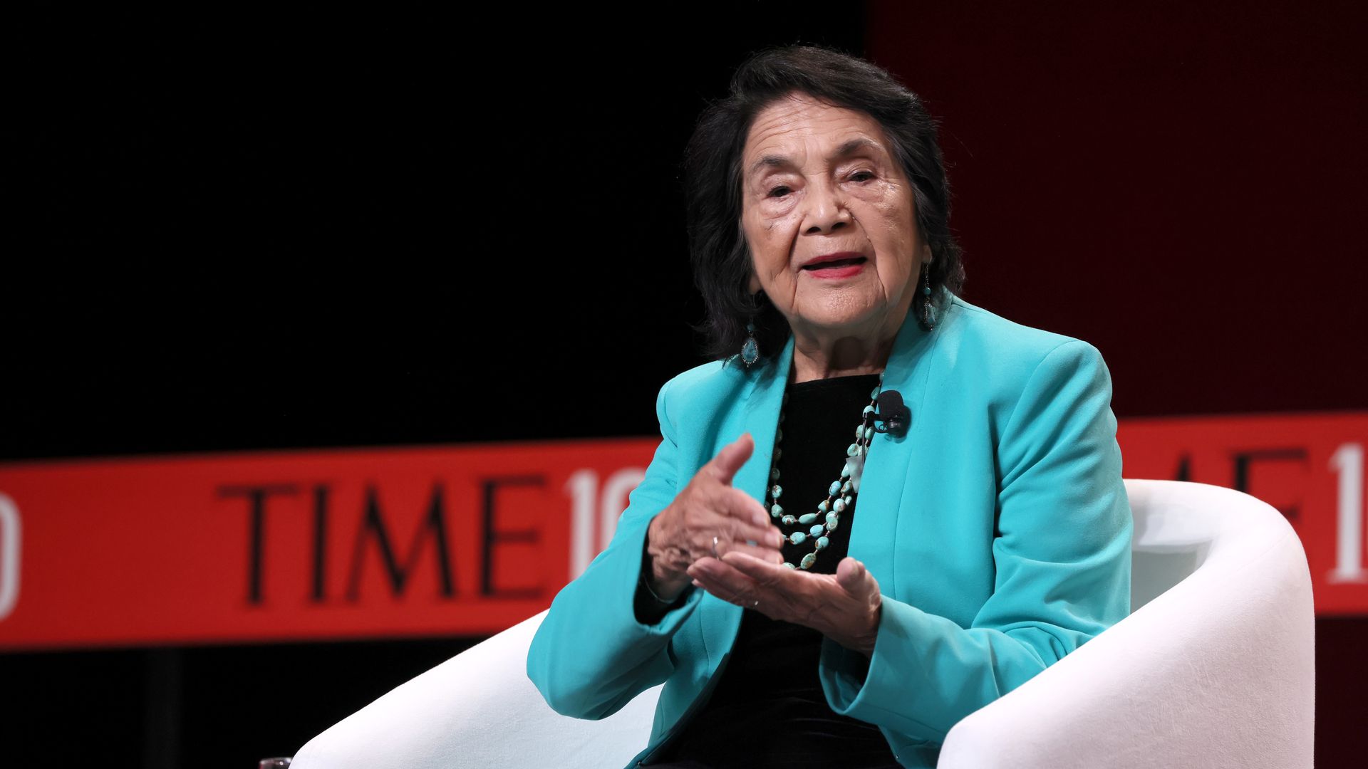 Photo of Dolores Huerta in a mint green blazer speaking from a white chair on stage at a Time 100 event
