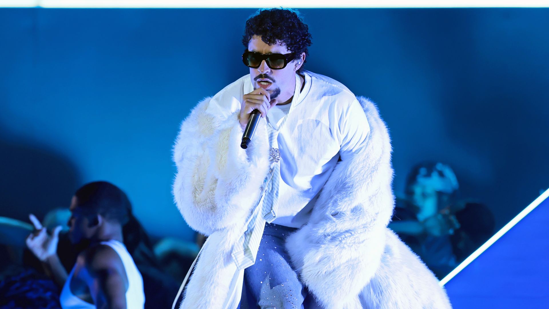 A photo of Bad Bunny on stage, holding a microphone. He's wear a white coat and a white shirt with sunglasses.