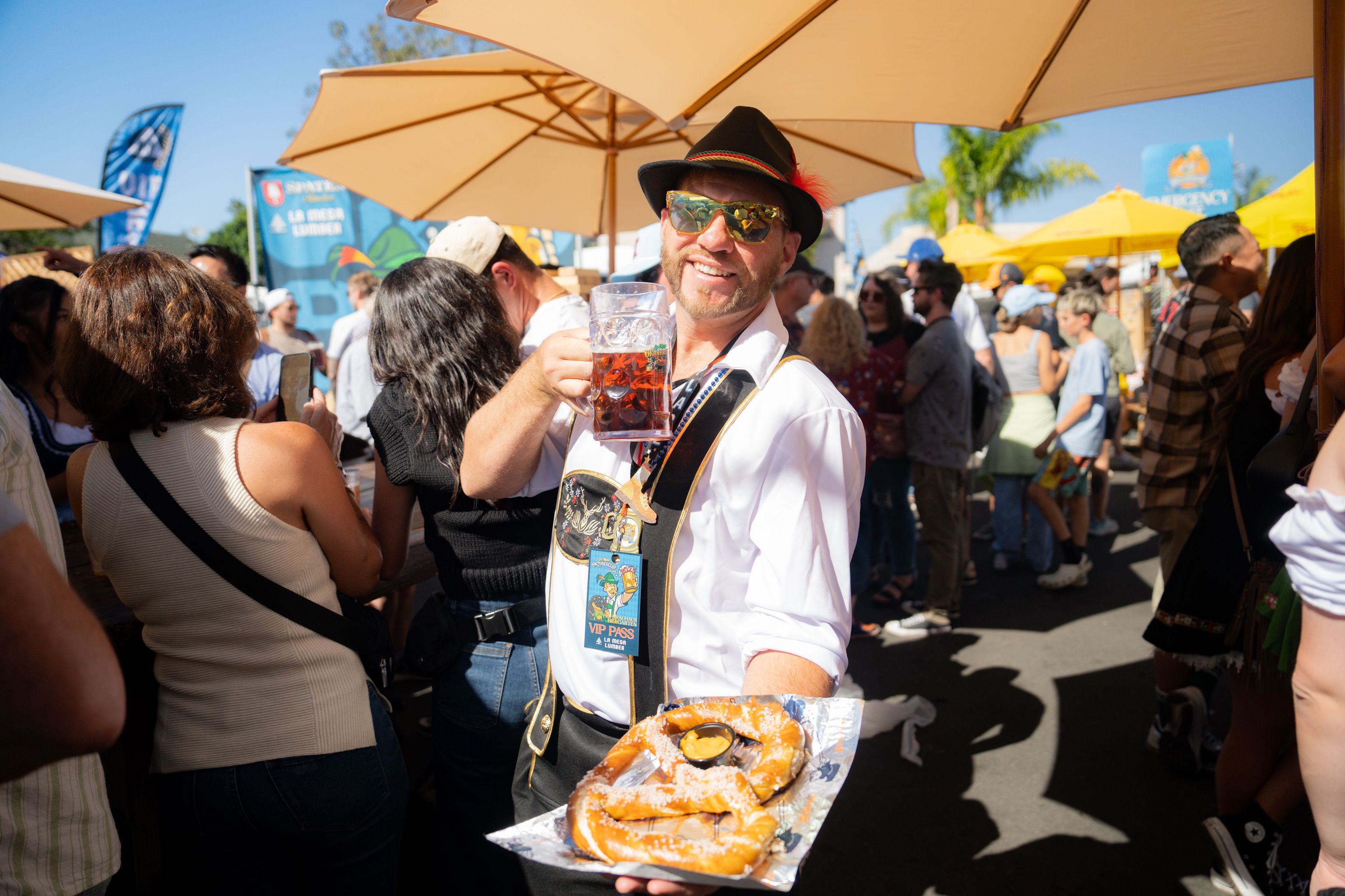 Smiling man in a black hat with a red feather and sunglasses holds a beer mug at a sunny outdoor festival. A pretzel on a plate sits in the foreground; crowd with yellow umbrellas and banners behind.