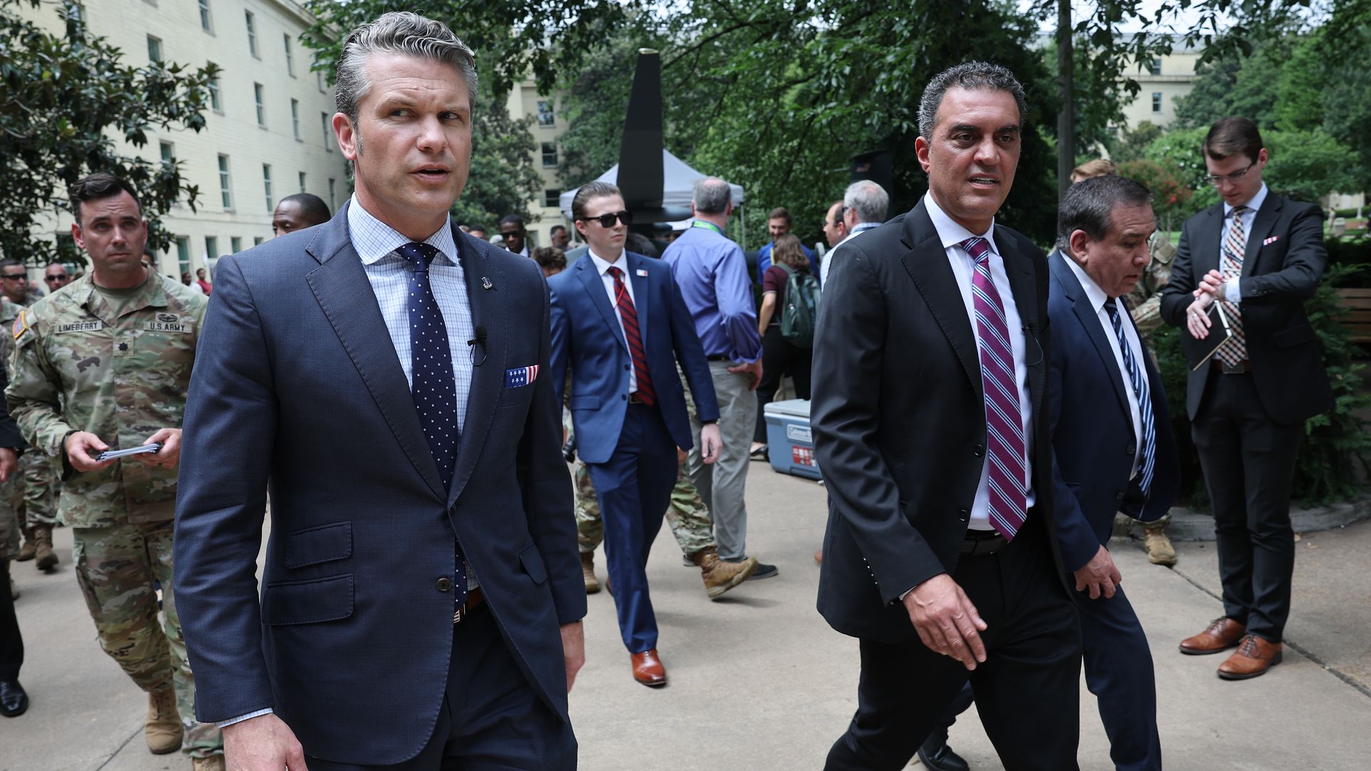 Group of men in suits walking outdoors; foreground man in a navy suit, others in business attire, with soldiers in camouflage and green trees in the background.