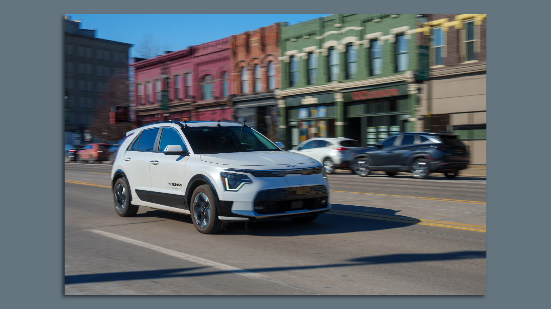 Image of a white Kia Niro EV, with Corktown Carshare on the side, driving along Michigan Avenue in Detroit. 